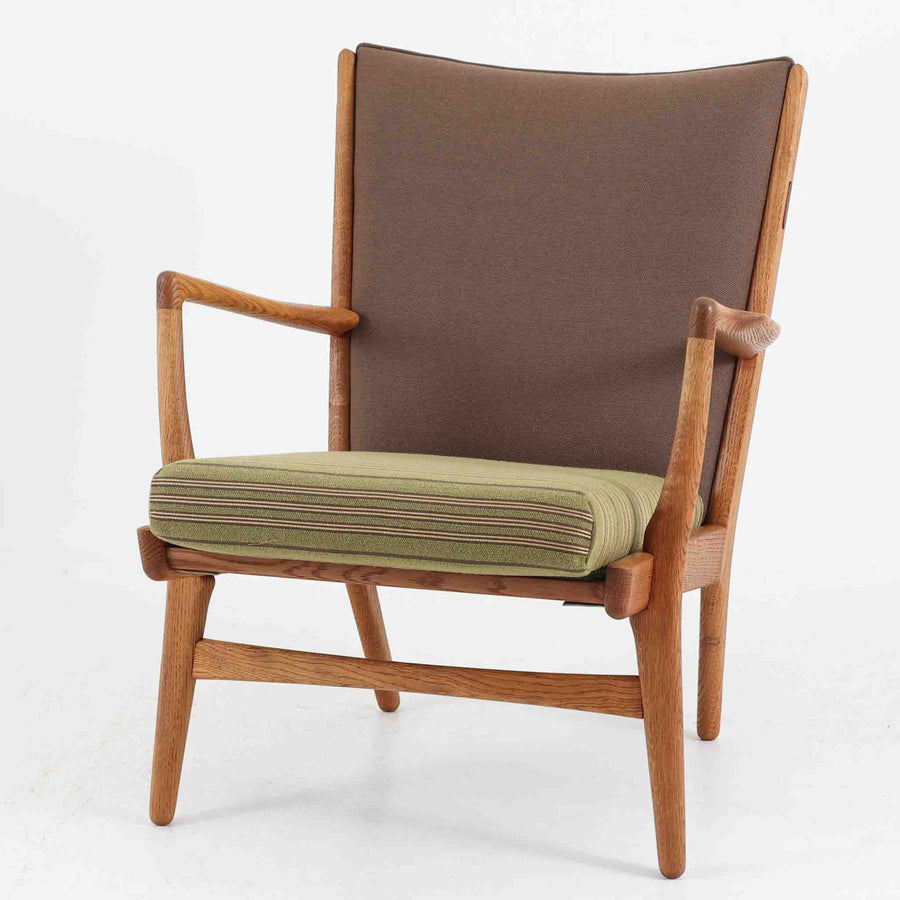 Hans J. Wegner（ハンス・J・ウェグナー）によるイージーチェア「AP16」。デンマークの名門AP stolen社より製造された一脚です。緩やかに傾斜した背もたれと、滑らかに削り出されたアームの曲線が美しく調和し、オーク材の力強い木目と職人技が際立ちます。背面にはブラウン、座面にはグリーンストライプの張地を採用し、上品ながらも温かみのある印象を与えます。現地デンマークのオークションで入手した確かな出自を持つ本品は、読書やくつろぎの時間をより豊かなものにしてくれます。経年による木部の風合いがさらに深みを加えています。
