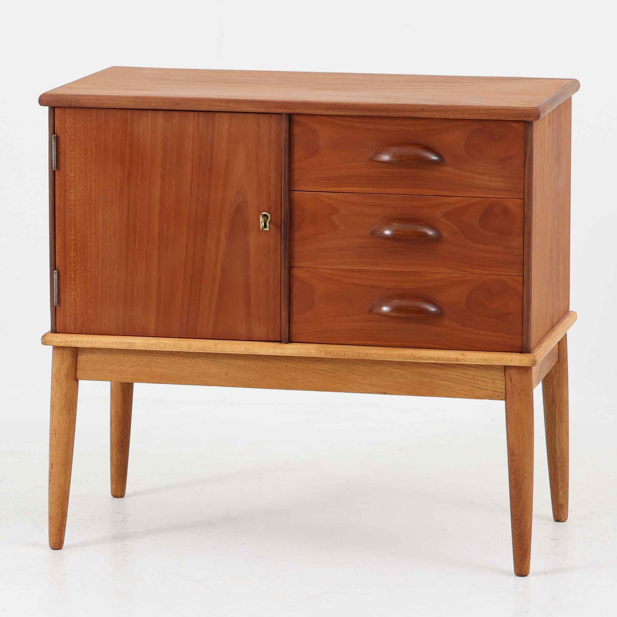 デンマーク製の小ぶりなキャビネットです。チーク材をメインに、オーク材やビーチ材を組み合わせることで、温かみのある色調と軽快な雰囲気を併せ持っています。左側は扉付き収納、右側には3杯の引き出しを備え、実用性とデザイン性を両立。丸みを帯びた取っ手や脚部のテーパーラインが北欧家具らしい柔らかな印象を与えます。幅63cmとコンパクトなサイズで、リビングや寝室、玄関など限られたスペースでも活躍する、ビンテージならではの風合いを持つ一台です。