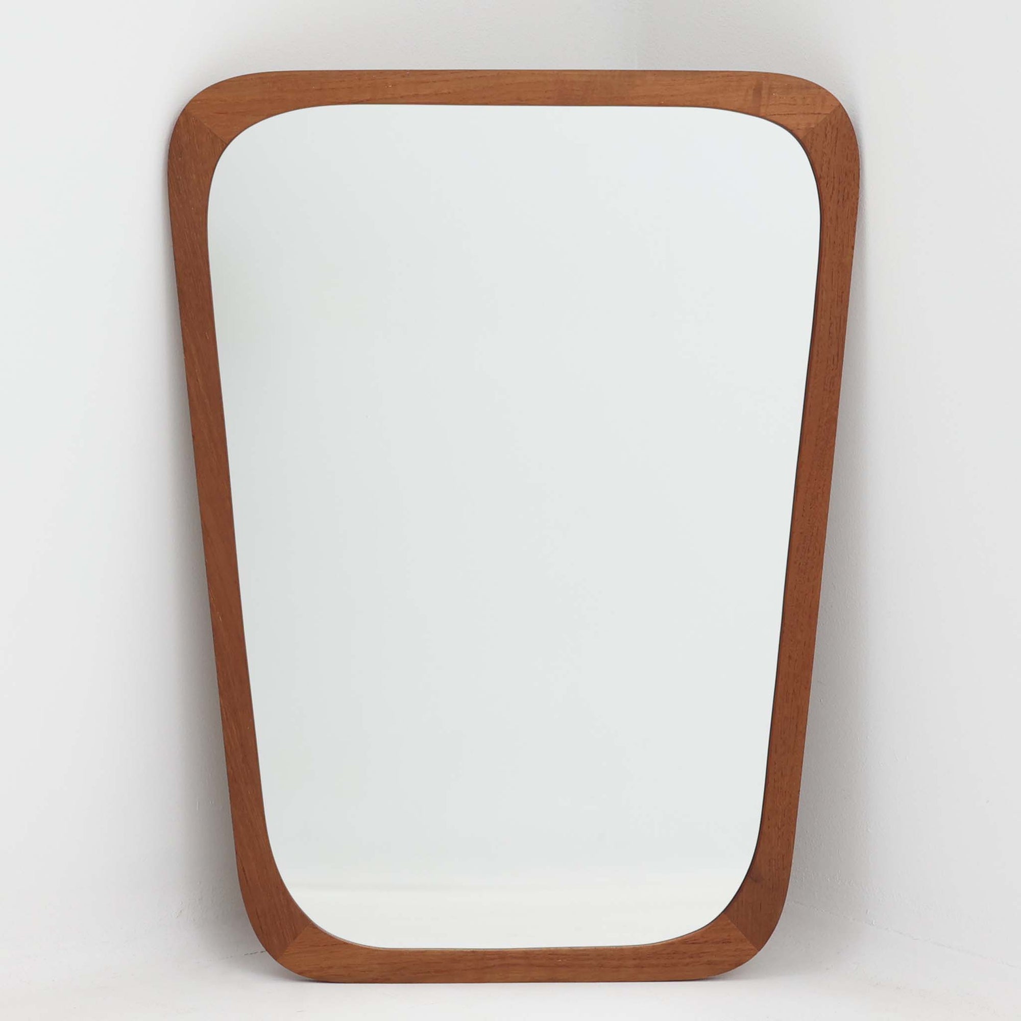 デンマークのAarhus Glasimport og Glassliberi社製による、チーク材フレームの壁掛け／立掛けミラーです。上部がわずかに広がる特徴的な台形フォルムが印象的で、やわらかな曲線を描くフレームが北欧デザインらしい優美さを感じさせます。チーク材のあたたかみのある色合いと美しい木目が空間に自然と溶け込み、玄関や寝室、リビングなどさまざまなシーンで活躍します。経年により深みを増した風合いが、北欧ビンテージらしい落ち着いた存在感を放つ一品です。