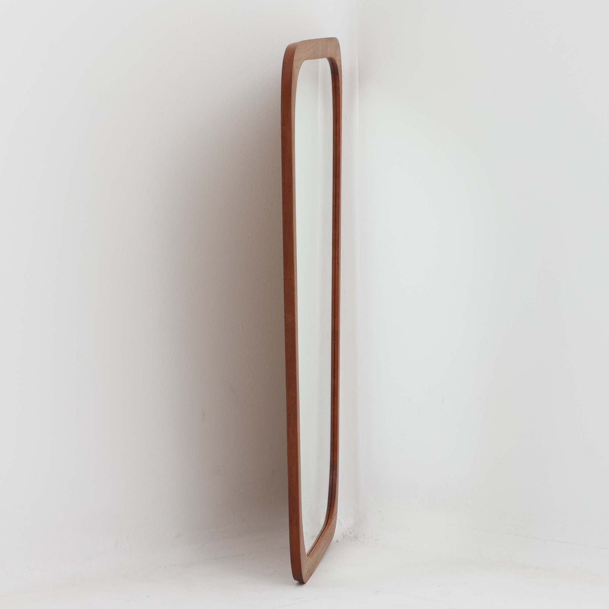 デンマークのAarhus Glasimport og Glassliberi社製による、チーク材フレームの壁掛け／立掛けミラーです。上部がわずかに広がる特徴的な台形フォルムが印象的で、やわらかな曲線を描くフレームが北欧デザインらしい優美さを感じさせます。チーク材のあたたかみのある色合いと美しい木目が空間に自然と溶け込み、玄関や寝室、リビングなどさまざまなシーンで活躍します。経年により深みを増した風合いが、北欧ビンテージらしい落ち着いた存在感を放つ一品です。