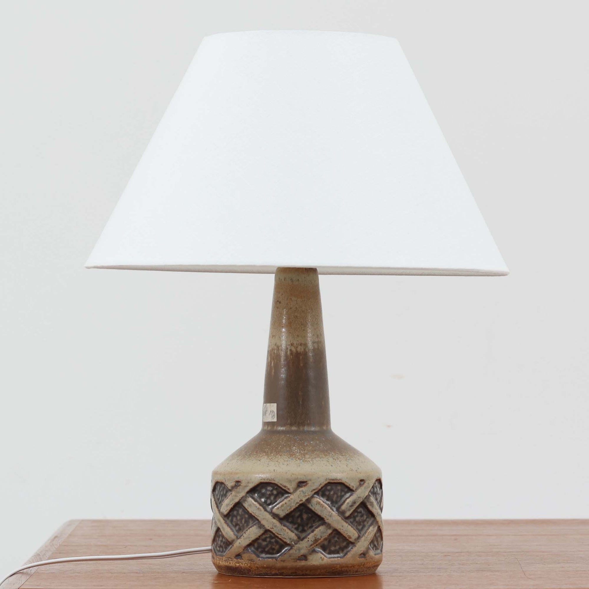 スーホルム・セラミック社製のこちらのテーブルランプは、デンマークで製造された北欧ビンテージ品で、高さ44cmと存在感のあるサイズ感が魅力です。陶磁器のベースには幾何学的なレリーフ模様が施され、釉薬の自然なグラデーションがビンテージらしい風合いを引き立てています。リネンシェードを通した柔らかな光は温かみを帯び、リビングやベッドサイドに心地よい雰囲気をもたらします。年月を経て深みを増した質感と、クラフトマンシップを感じさせる一点物ならではの魅力をお楽しみいただけます。
