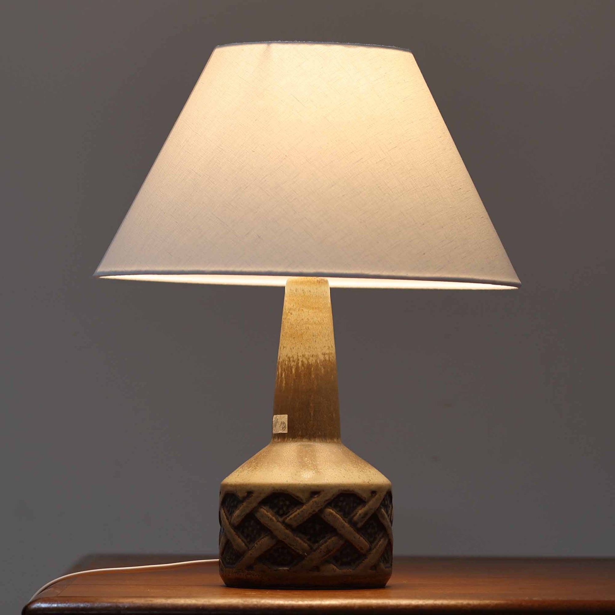 スーホルム・セラミック社製のこちらのテーブルランプは、デンマークで製造された北欧ビンテージ品で、高さ44cmと存在感のあるサイズ感が魅力です。陶磁器のベースには幾何学的なレリーフ模様が施され、釉薬の自然なグラデーションがビンテージらしい風合いを引き立てています。リネンシェードを通した柔らかな光は温かみを帯び、リビングやベッドサイドに心地よい雰囲気をもたらします。年月を経て深みを増した質感と、クラフトマンシップを感じさせる一点物ならではの魅力をお楽しみいただけます。