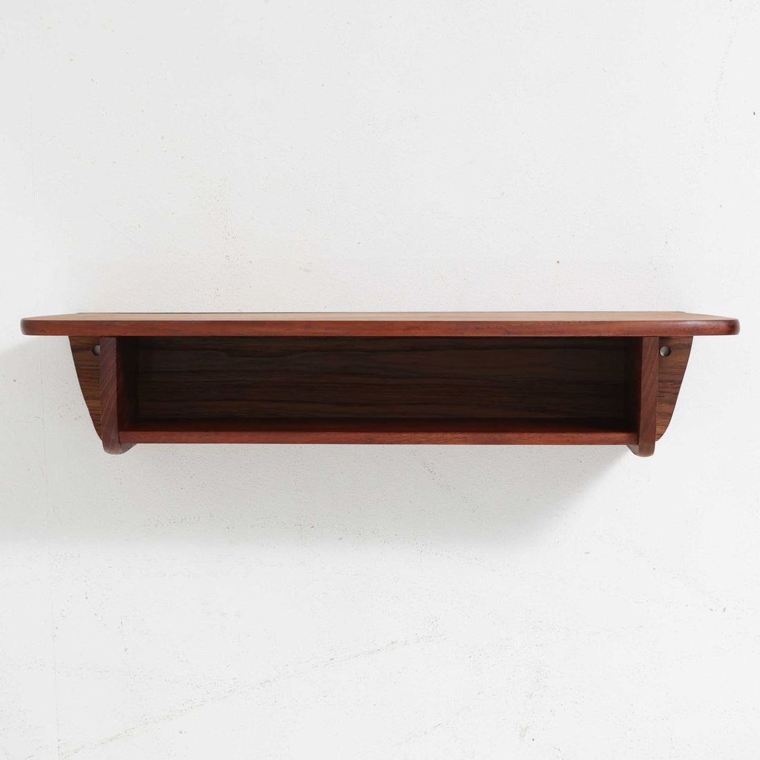 デンマーク製ウォールシェルフです。上質なチーク無垢材を使用し、滑らかなカーブを描く縁取りと丸みのあるフォルムが北欧家具らしい柔らかさを感じさせます。奥行きを抑えたコンパクトなサイズで、玄関や寝室、リビングのちょっとしたスペースにも取り付けやすい設計です。経年により深みを増したチーク材の美しい色合いが、壁面を上品に演出します。