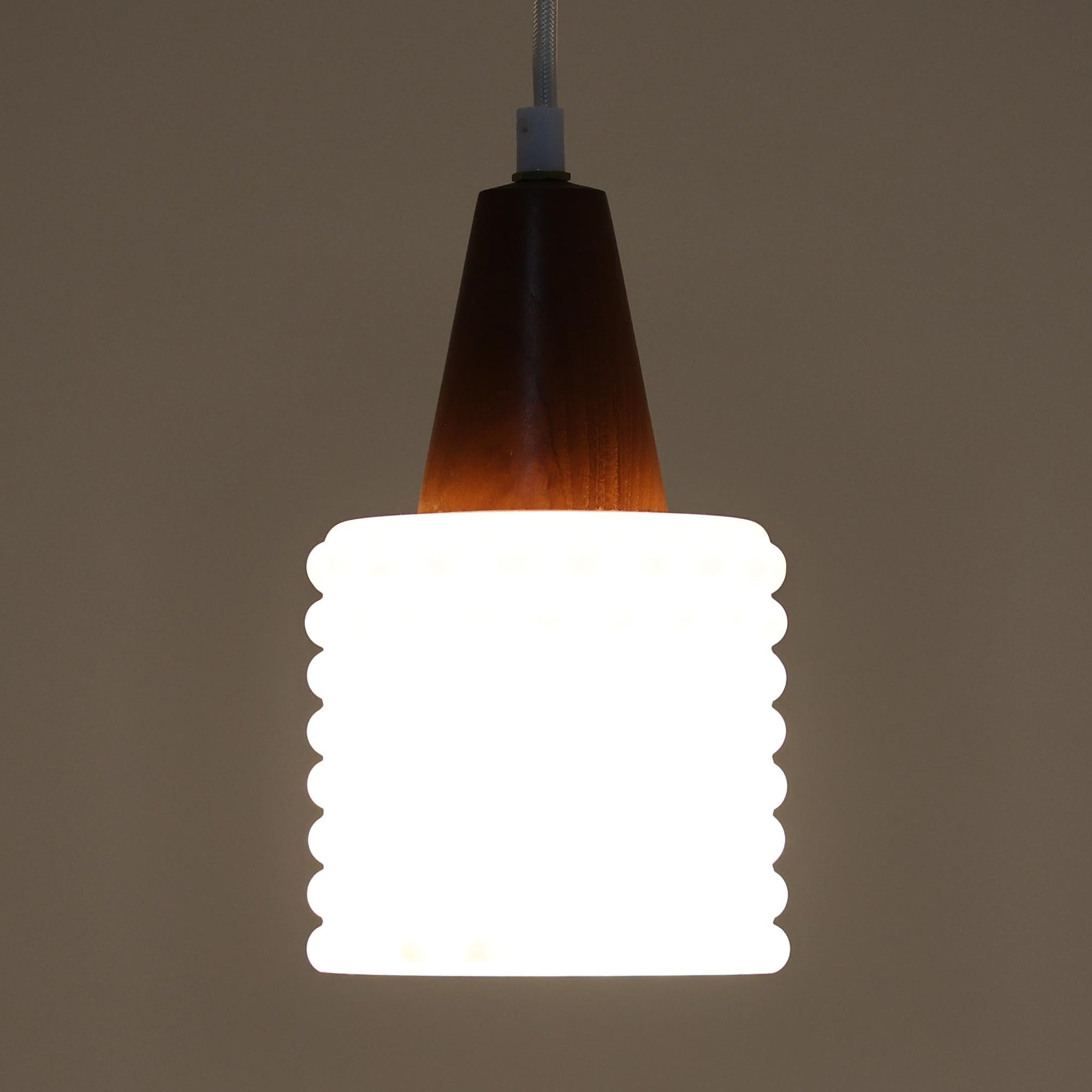 デンマーク製のペンダントライト、直径13cmです。ガラスシェードには立体的なドット模様が施され、点灯すると陰影が浮かび上がり独特の表情を生み出します。上部にはチーク材が用いられ、ガラスの繊細さと木の温もりが調和した北欧らしい意匠が魅力です。小ぶりなサイズながらも存在感があり、キッチンや廊下、ベッドサイドなど複数使いにも適しています。経年による素材の風合いがビンテージ品ならではの味わいを感じさせ、日常空間に個性を添える一品です。