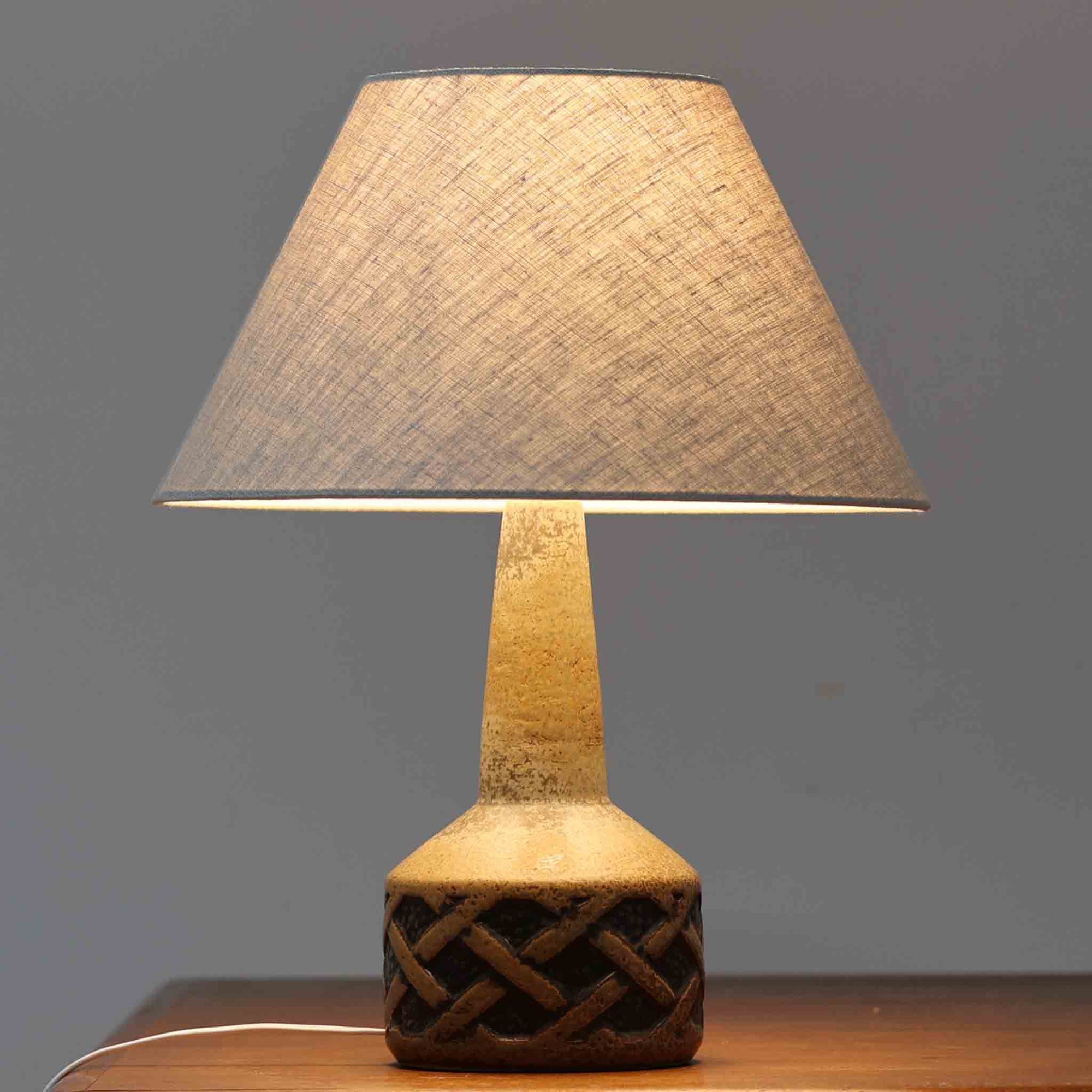 デンマークの名窯スーホルム・セラミック社製のこちらのテーブルランプは、高さ44cmと程よいサイズ感で、北欧ビンテージならではの存在感を放ちます。陶磁器製のベースには立体的な幾何学模様が施され、釉薬のグラデーションが経年によって深みを増し、独特の風合いを醸し出しています。リネンシェードから広がる柔らかな光は、リビングやベッドサイドに落ち着きある雰囲気を演出。シンプルでありながら温もりを感じさせる一点物の魅力をお楽しみいただけます。