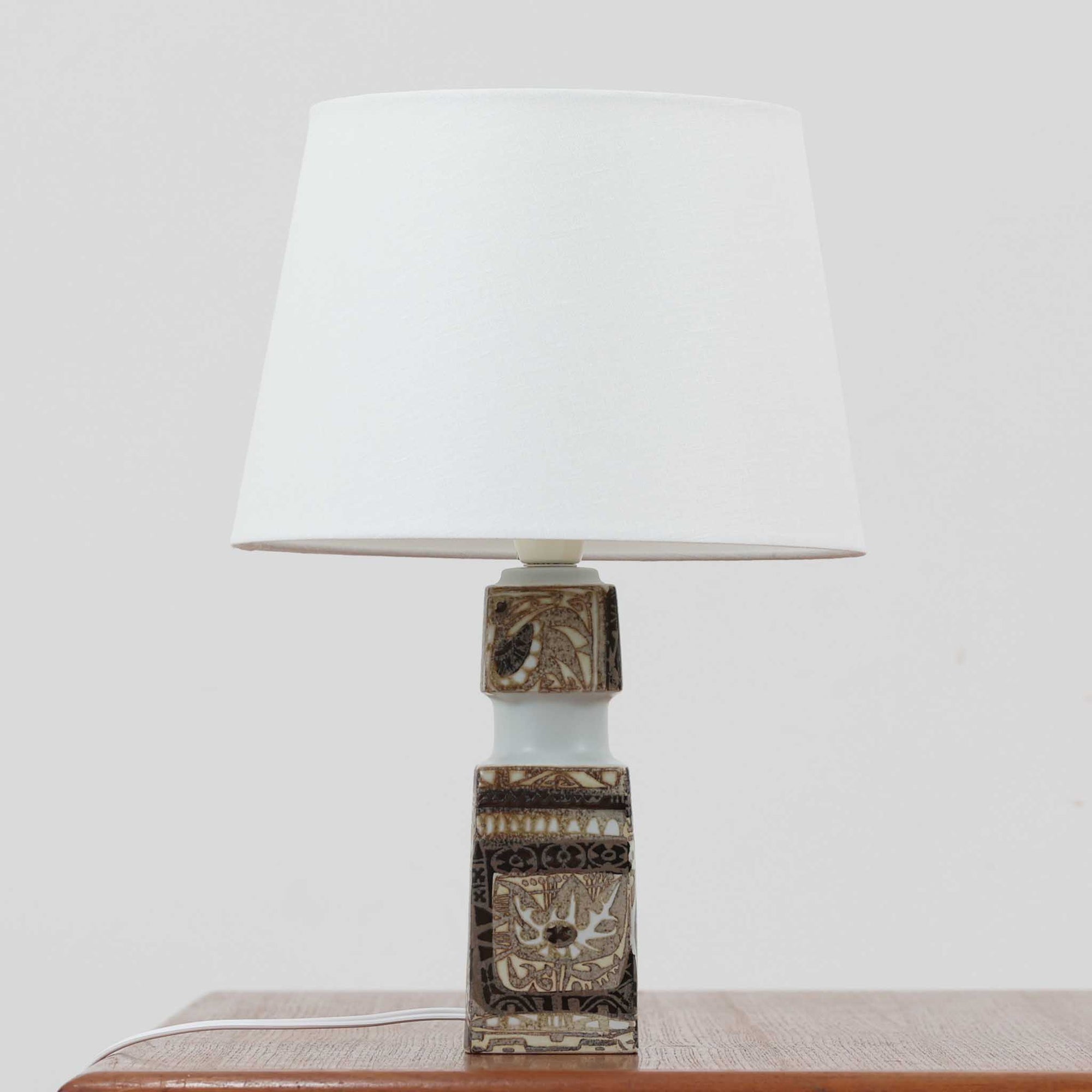 Nils Thorssonがデザインし、Royal Copenhagen社から発表された「BACA」シリーズのテーブルランプは、高さ39cmのバランスの取れたサイズ感で、北欧ビンテージらしい存在感を放ちます。陶磁器製のベースには細やかな装飾模様が施され、グラフィカルでありながら落ち着いた色調が空間に洗練された雰囲気を添えます。リネンシェードを透過する柔らかな光が温もりを演出し、リビングやベッドサイドに最適です。年月を経て深みを増した質感と工芸的な美しさを楽しめる一点物です。