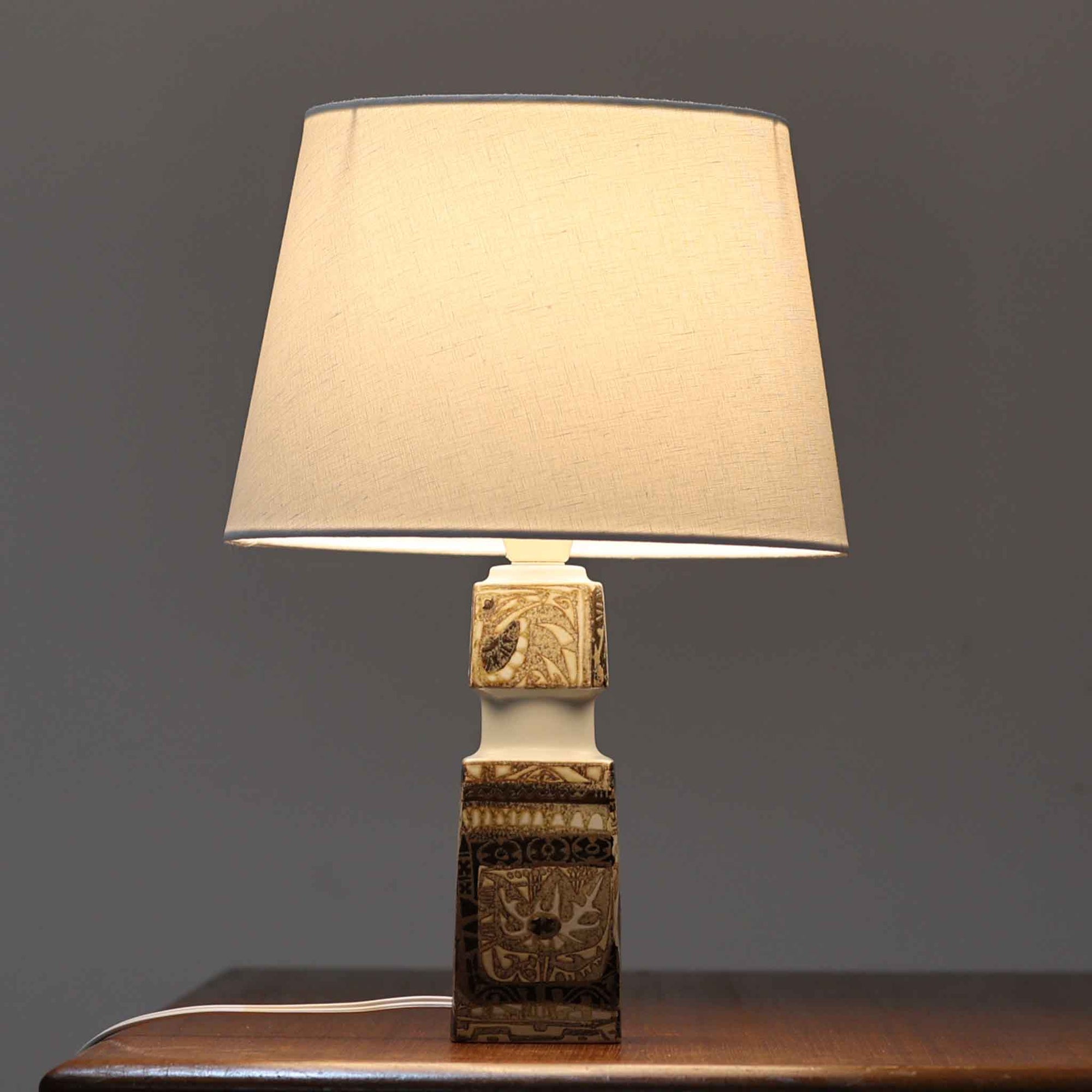 Nils Thorssonがデザインし、Royal Copenhagen社から発表された「BACA」シリーズのテーブルランプは、高さ39cmのバランスの取れたサイズ感で、北欧ビンテージらしい存在感を放ちます。陶磁器製のベースには細やかな装飾模様が施され、グラフィカルでありながら落ち着いた色調が空間に洗練された雰囲気を添えます。リネンシェードを透過する柔らかな光が温もりを演出し、リビングやベッドサイドに最適です。年月を経て深みを増した質感と工芸的な美しさを楽しめる一点物です。