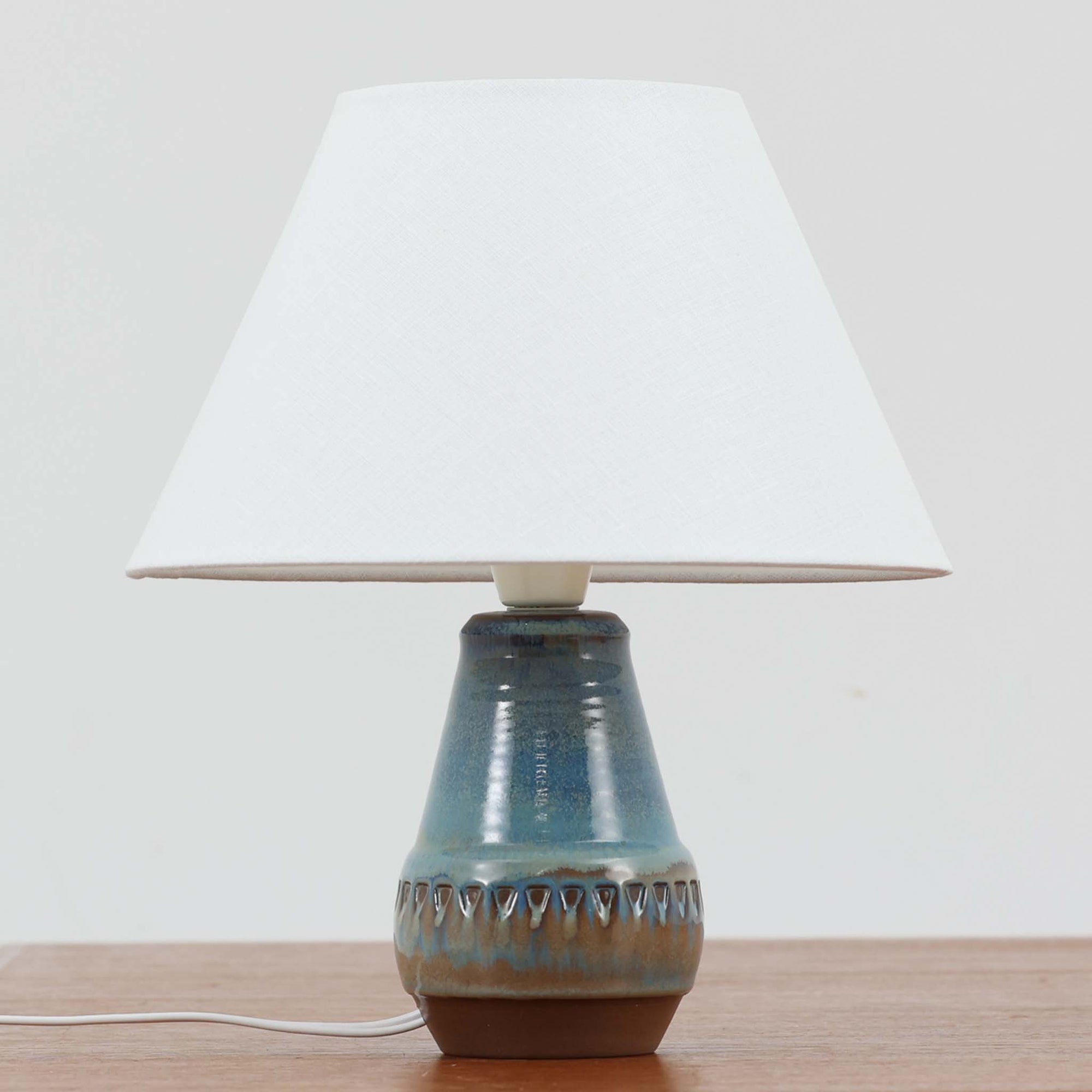 デンマークの名窯Michael Andersen & Sonによる高さ29cmのテーブルランプは、北欧ビンテージらしい温もりと存在感を兼ね備えています。陶磁器製のベースにはブルーからブラウンへと移ろう釉薬が美しいグラデーションを描き、下部に施された幾何学的なレリーフ模様がデザインのアクセントとなっています。リネンシェードを透過する柔らかな光は、ベッドサイドやリビングに穏やかな雰囲気を添え、使うほどに味わいを深める一点物の魅力を楽しめます。