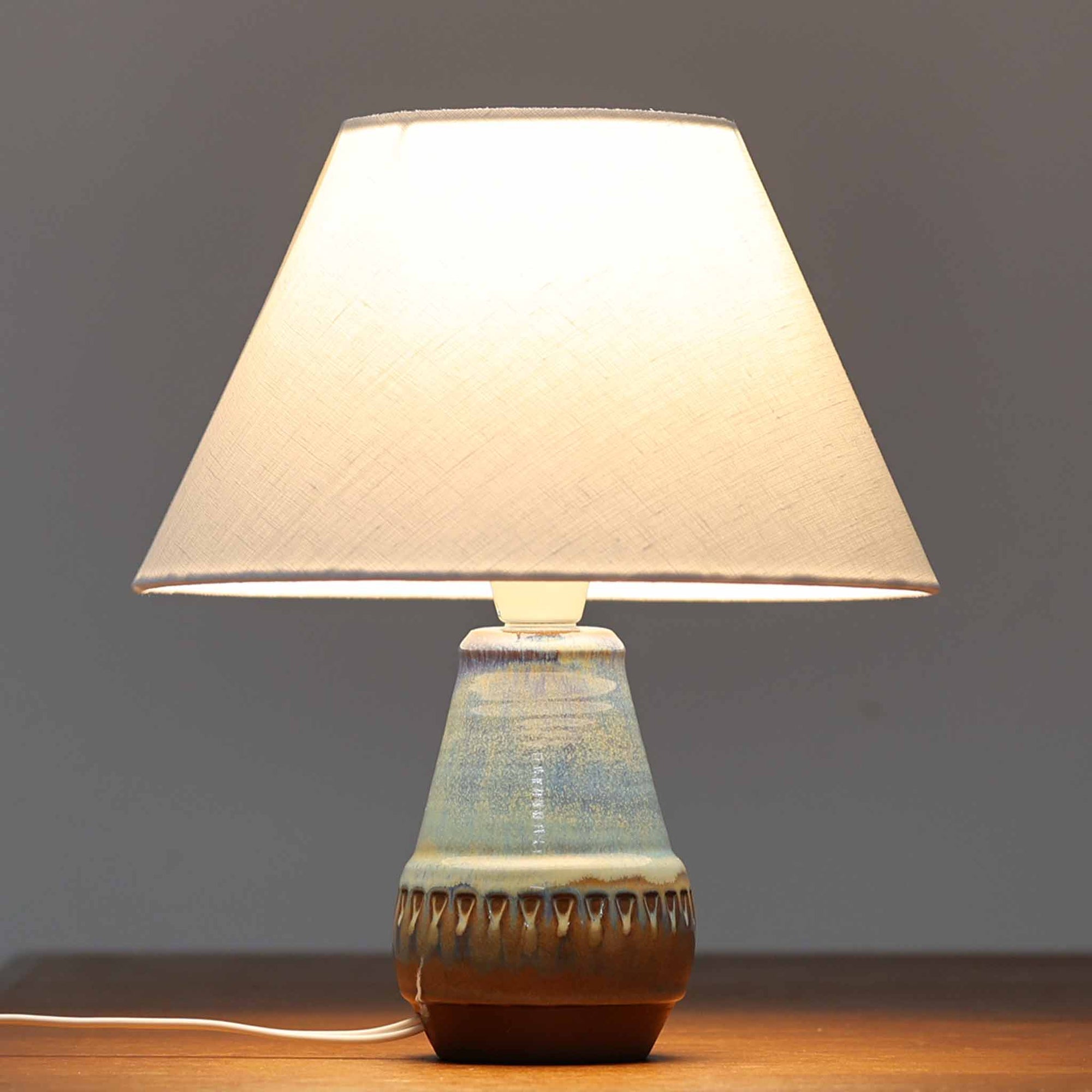 デンマークの名窯Michael Andersen & Sonによる高さ29cmのテーブルランプは、北欧ビンテージらしい温もりと存在感を兼ね備えています。陶磁器製のベースにはブルーからブラウンへと移ろう釉薬が美しいグラデーションを描き、下部に施された幾何学的なレリーフ模様がデザインのアクセントとなっています。リネンシェードを透過する柔らかな光は、ベッドサイドやリビングに穏やかな雰囲気を添え、使うほどに味わいを深める一点物の魅力を楽しめます。