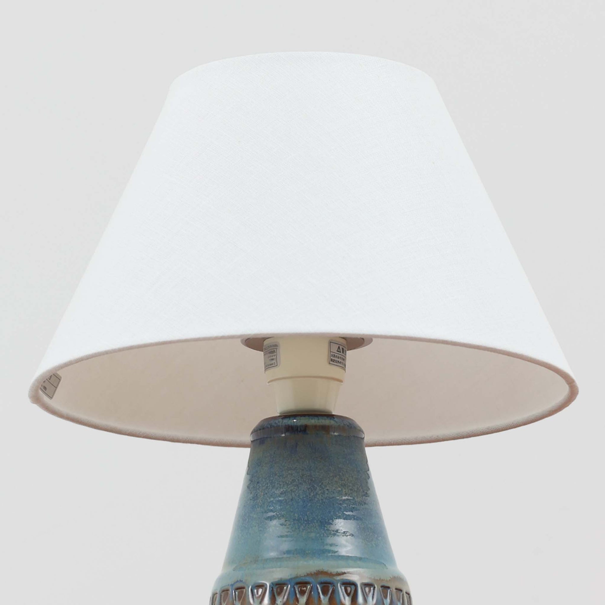 デンマークの名窯Michael Andersen &amp; Sonによる高さ29cmのテーブルランプは、北欧ビンテージらしい温もりと存在感を兼ね備えています。陶磁器製のベースにはブルーからブラウンへと移ろう釉薬が美しいグラデーションを描き、下部に施された幾何学的なレリーフ模様がデザインのアクセントとなっています。リネンシェードを透過する柔らかな光は、ベッドサイドやリビングに穏やかな雰囲気を添え、使うほどに味わいを深める一点物の魅力を楽しめます。