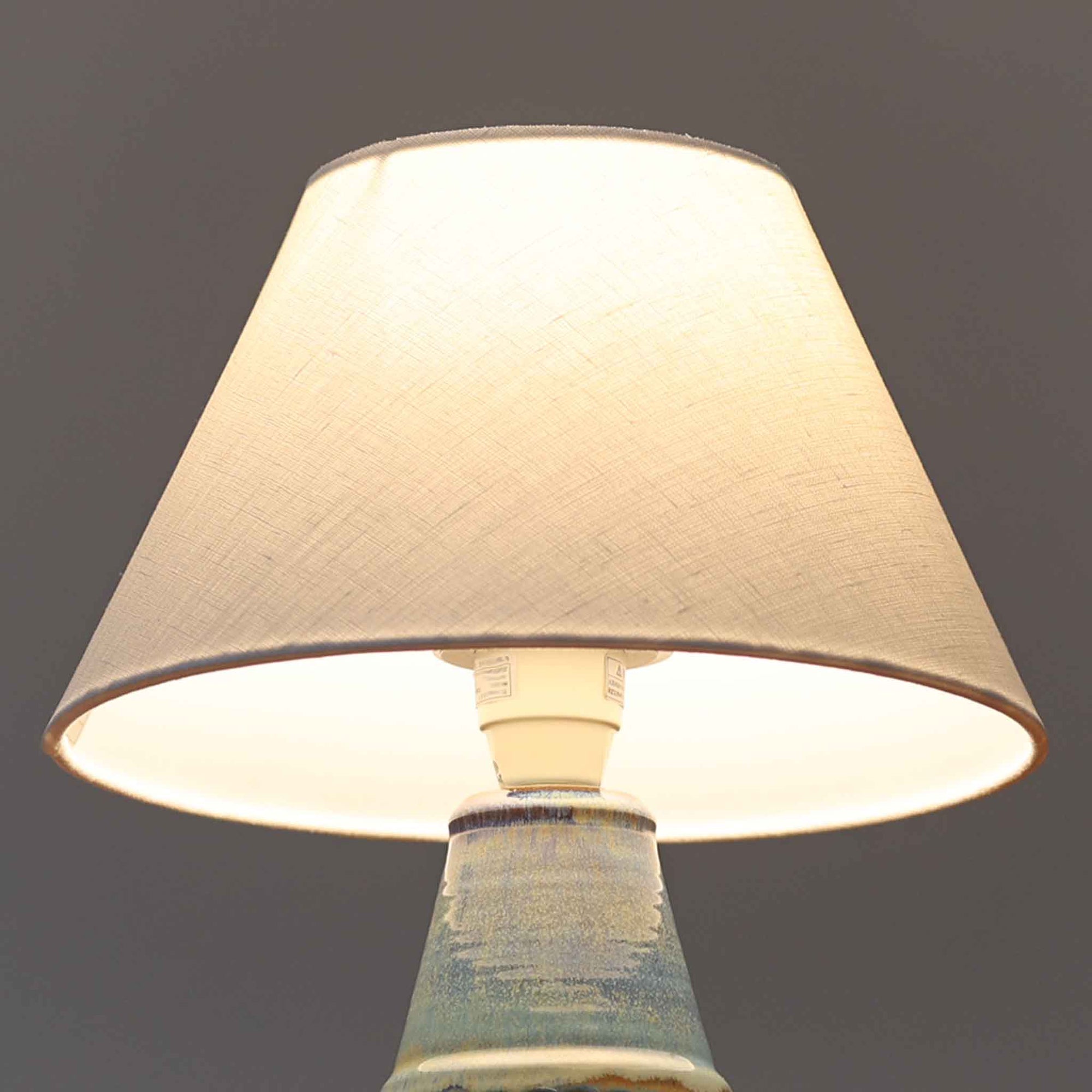 デンマークの名窯Michael Andersen &amp; Sonによる高さ29cmのテーブルランプは、北欧ビンテージらしい温もりと存在感を兼ね備えています。陶磁器製のベースにはブルーからブラウンへと移ろう釉薬が美しいグラデーションを描き、下部に施された幾何学的なレリーフ模様がデザインのアクセントとなっています。リネンシェードを透過する柔らかな光は、ベッドサイドやリビングに穏やかな雰囲気を添え、使うほどに味わいを深める一点物の魅力を楽しめます。