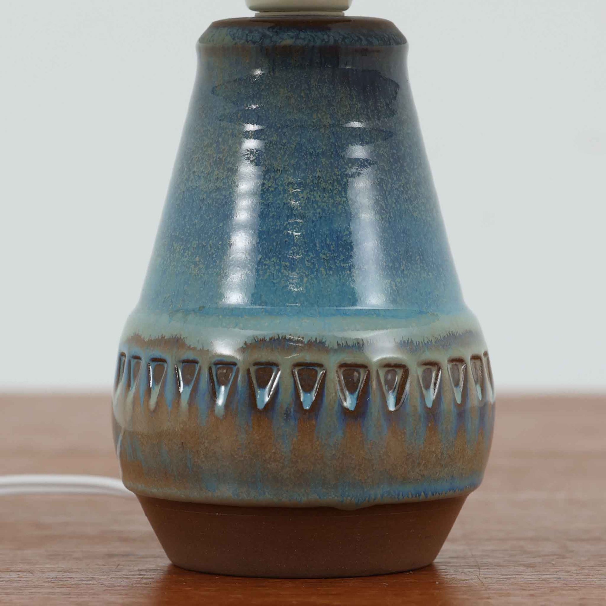 デンマークの名窯Michael Andersen &amp; Sonによる高さ29cmのテーブルランプは、北欧ビンテージらしい温もりと存在感を兼ね備えています。陶磁器製のベースにはブルーからブラウンへと移ろう釉薬が美しいグラデーションを描き、下部に施された幾何学的なレリーフ模様がデザインのアクセントとなっています。リネンシェードを透過する柔らかな光は、ベッドサイドやリビングに穏やかな雰囲気を添え、使うほどに味わいを深める一点物の魅力を楽しめます。