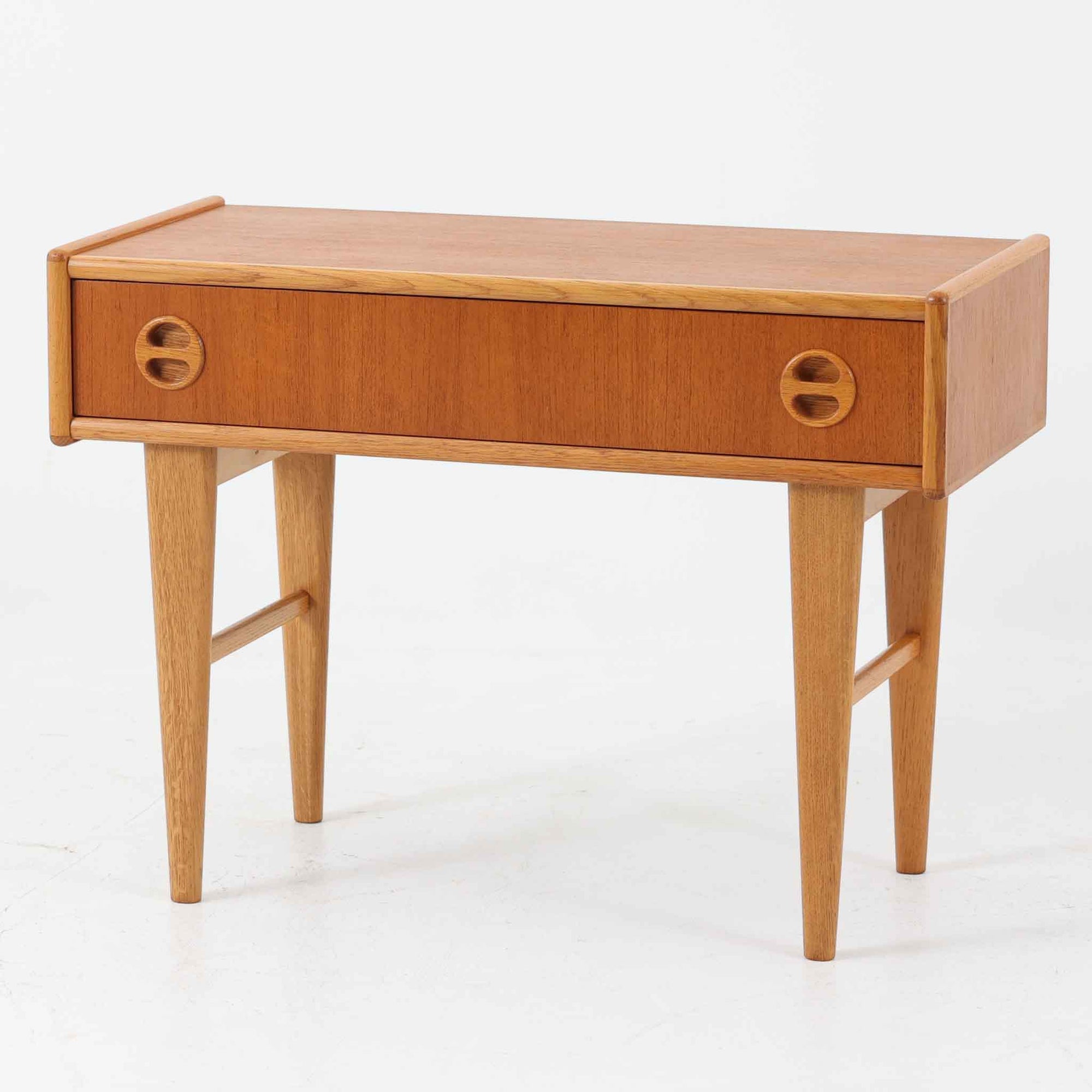 チーク材とオーク材を組み合わせたこちらの2段チェストは、幅70cmのコンパクトなサイズながら、北欧ビンテージ家具らしい洗練されたデザインが魅力です。丸型の埋め込み取手が愛らしいアクセントとなり、テーパー脚との調和が軽快な印象を与えます。引き出しは小物収納に適しており、ベッドサイドやリビングのサイドテーブルとしても活躍。素材のコントラストと経年による木肌の深みが、空間に温もりと個性を添える一品です。