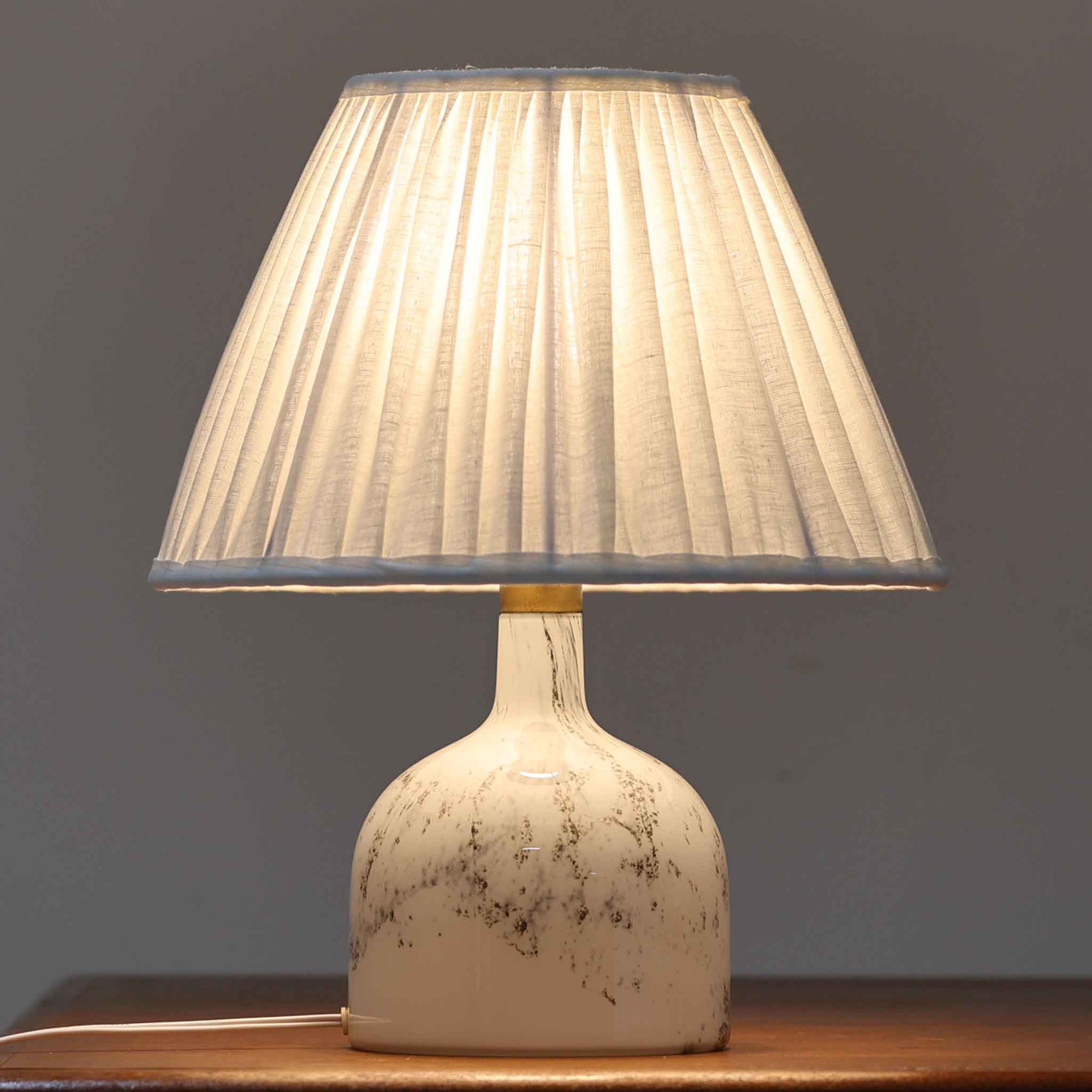 デンマークを代表するガラスメーカー、Holmegaard社よりMichael Bangが手掛けた「LAMP ART」テーブルランプは、高さ40cmのバランス良いサイズ感で、北欧ビンテージの魅力を放ちます。ベースはホワイトガラスにマーブル状の模様が施され、柔らかな光を映し出す造形美が特徴です。真鍮のパーツがアクセントとなり、リネンシェードを透過する光は空間に穏やかな温もりを添えます。洗練されたデザインと職人技が調和した一点物で、リビングや寝室の雰囲気を格上げする逸品です。