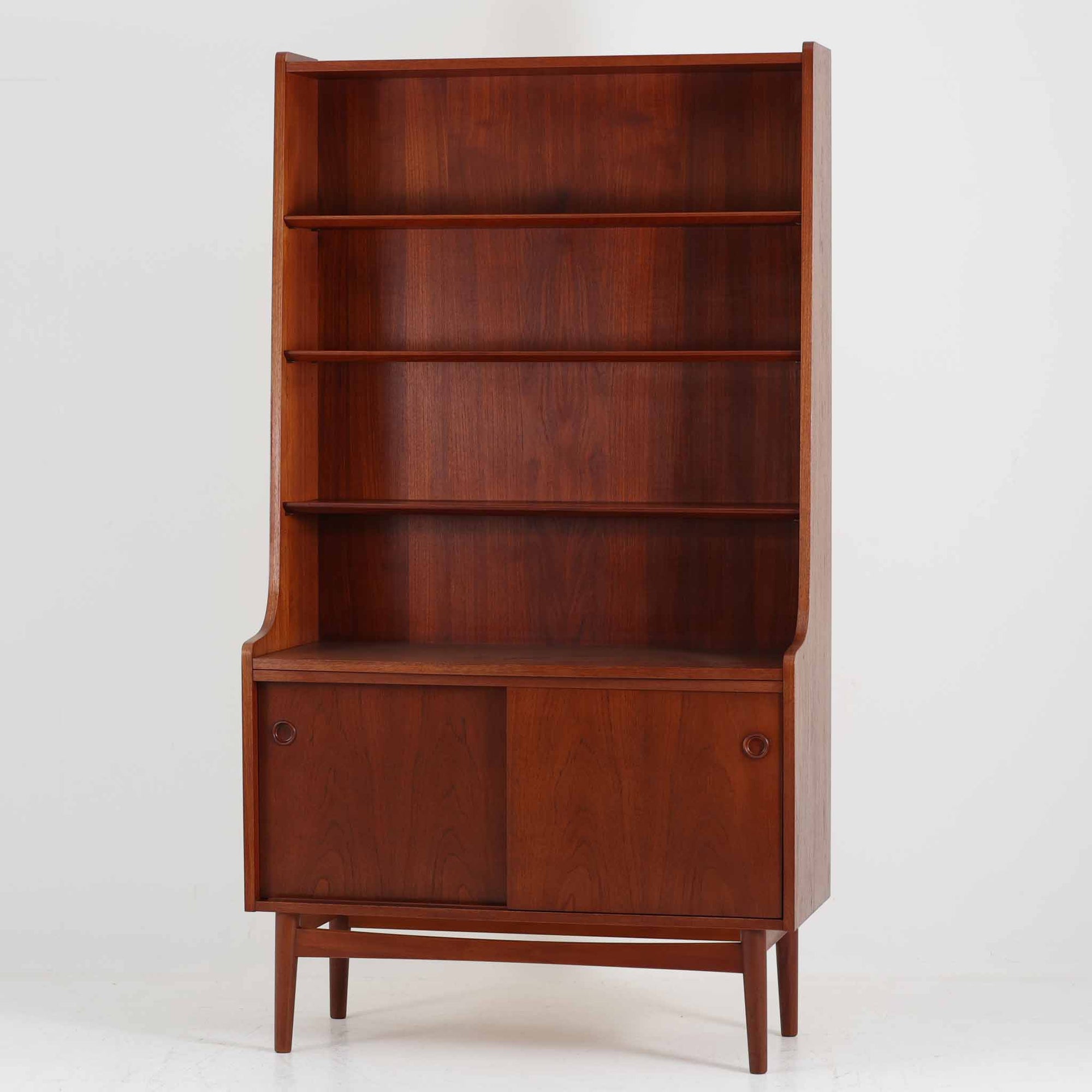 Johannes Sorth（ヨハネス・ソース）によるデザインで、Nexo Mobelfabrik社より製造されたチーク材のディスプレイシェルフです。上部の棚は本やオブジェを美しく飾ることができ、下部には引き戸収納を備え、実用性にも優れています。緩やかな曲線を描く側面のデザインが柔らかな印象を与え、北欧家具らしい温もりと機能美が感じられる一台です。脚部の細身のフォルムが軽やかさを演出し、リビングや書斎、ダイニングなど幅広い空間に調和します。