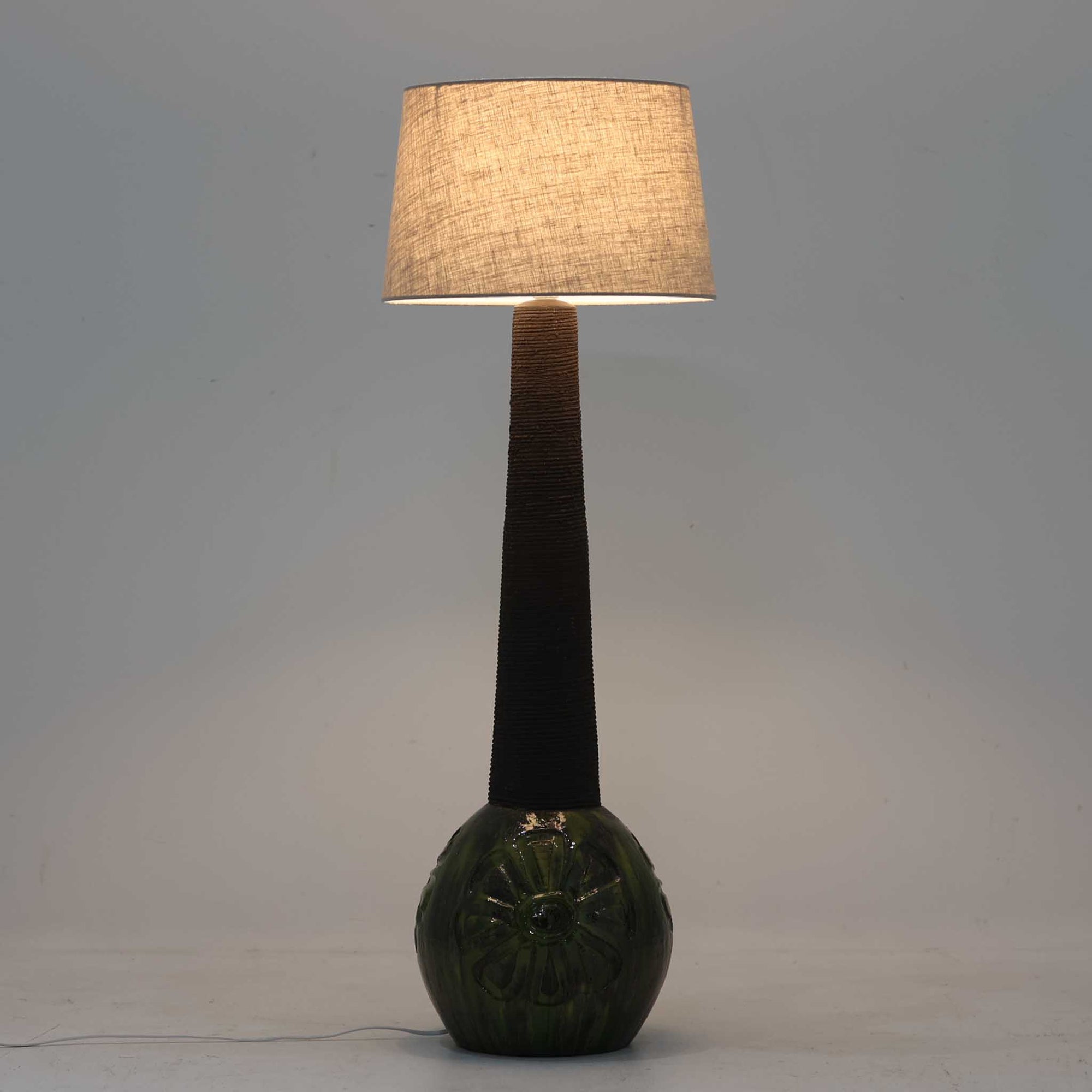 Soholm Keramik（スーホルム・セラミック）製による高さ95cmのビンテージランプです。ベースには緑色の釉薬で仕上げられた陶磁器が用いられ、花模様のレリーフが浮かび上がる温かみのあるデザインが魅力です。細身で長く伸びた支柱部分には素朴な質感が活かされ、上部にはシンプルなリネンシェードを合わせることで、柔らかな光を優しく拡散します。陶器製ランプとしては稀に見る大きさで、テーブルランプでありながらフロアランプのような存在感を放ち、リビングのアクセントや間接照明としても活躍します。陶芸的な味わいと経年の風合いが調和した、北欧ビンテージならではの逸品です。