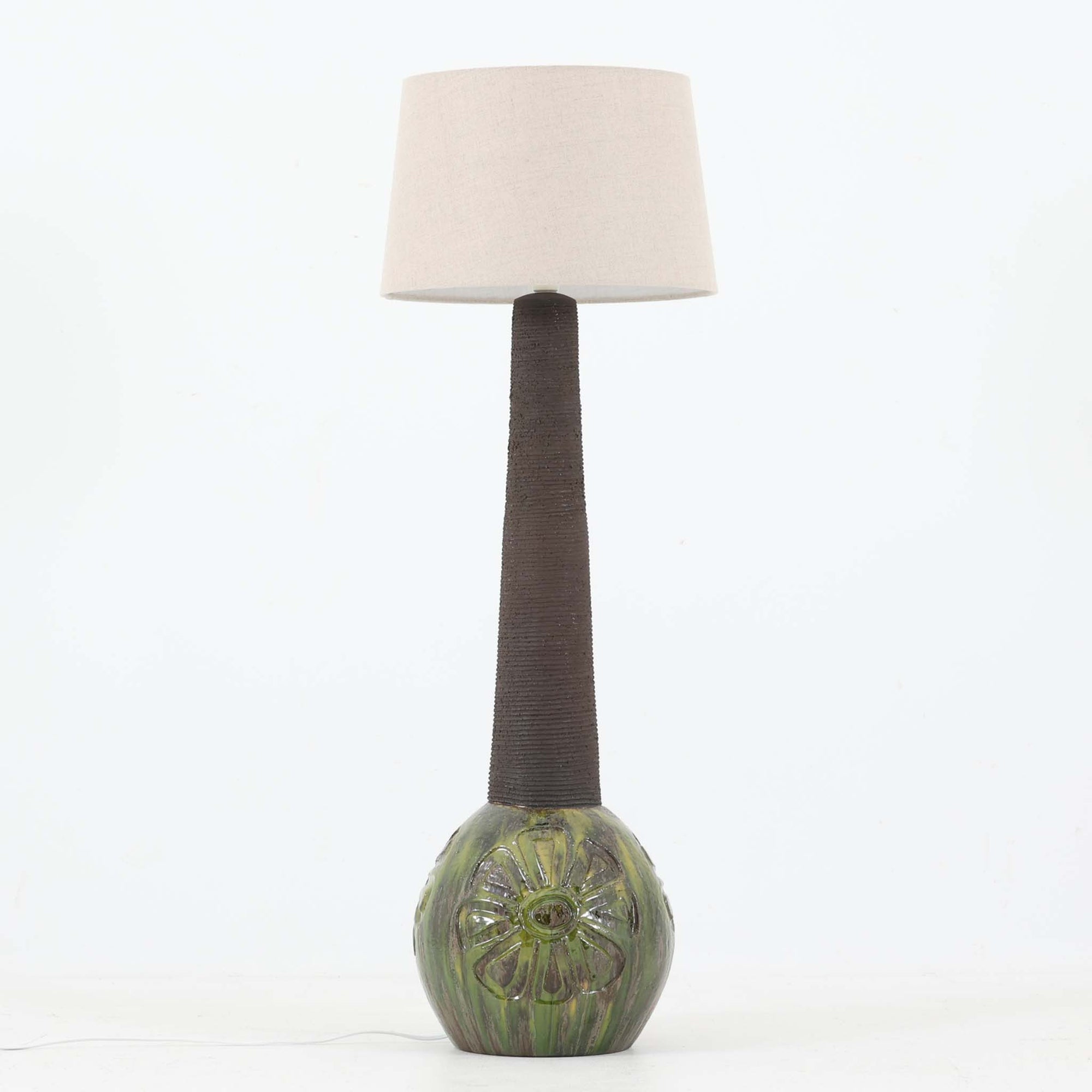 Soholm Keramik（スーホルム・セラミック）製による高さ95cmのビンテージランプです。ベースには緑色の釉薬で仕上げられた陶磁器が用いられ、花模様のレリーフが浮かび上がる温かみのあるデザインが魅力です。細身で長く伸びた支柱部分には素朴な質感が活かされ、上部にはシンプルなリネンシェードを合わせることで、柔らかな光を優しく拡散します。陶器製ランプとしては稀に見る大きさで、テーブルランプでありながらフロアランプのような存在感を放ち、リビングのアクセントや間接照明としても活躍します。陶芸的な味わいと経年の風合いが調和した、北欧ビンテージならではの逸品です。