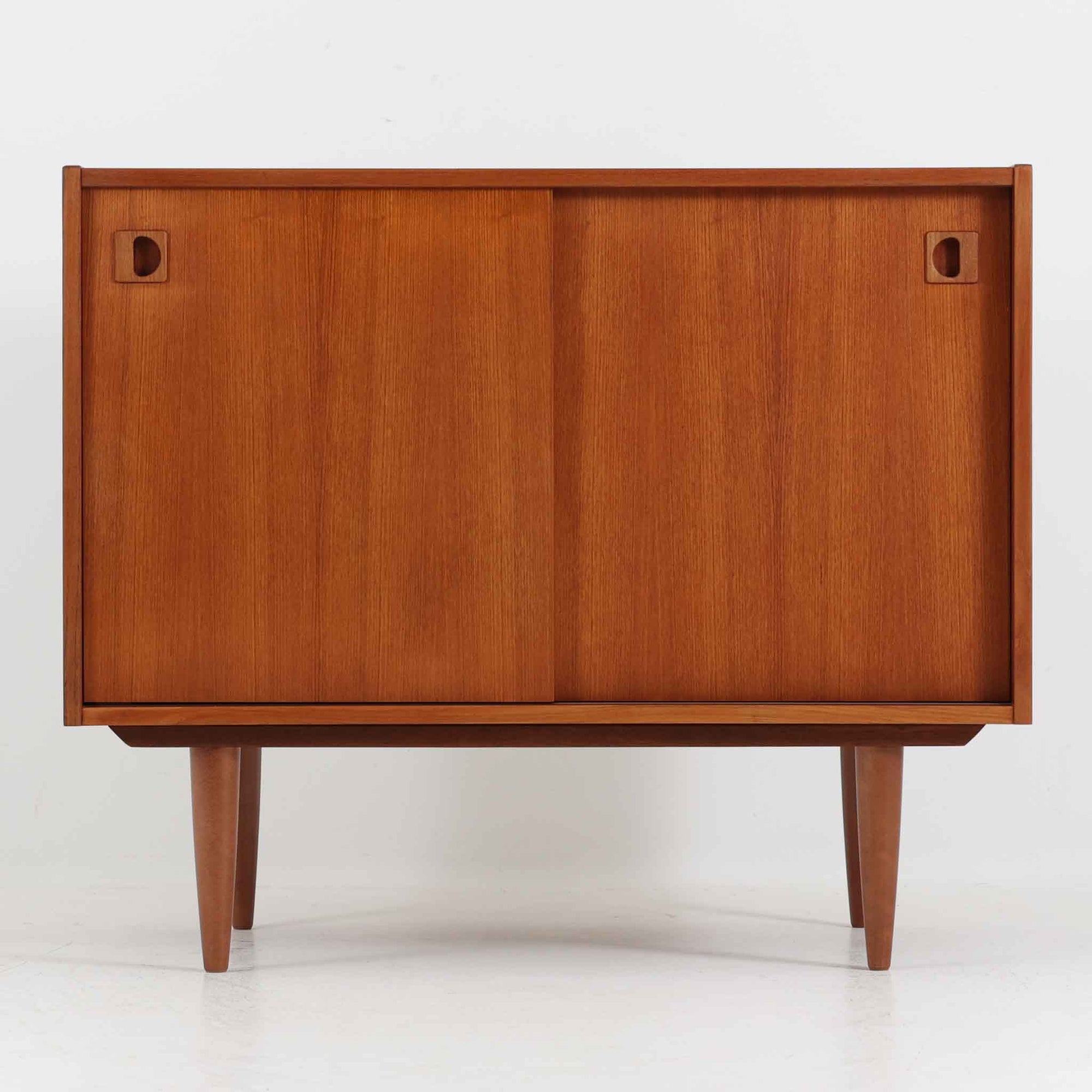 こちらの小ぶりなボードは、温かみのあるチーク材を用いたすっきりとした佇まいが魅力の北欧ビンテージ家具で、スライド扉を開くと左右に棚板付きの収納スペースが現れ、日用品や小物を効率よく整理できます。直線的な箱型の構造に、細身でテーパーの効いた脚部が軽やかな印象を添え、コンパクトながら存在感のある設計が空間に落ち着いたアクセントをもたらします。経年で深まった木肌の質感も味わい深く、リビングや玄関脇のサイド収納としても取り入れやすい一台です。