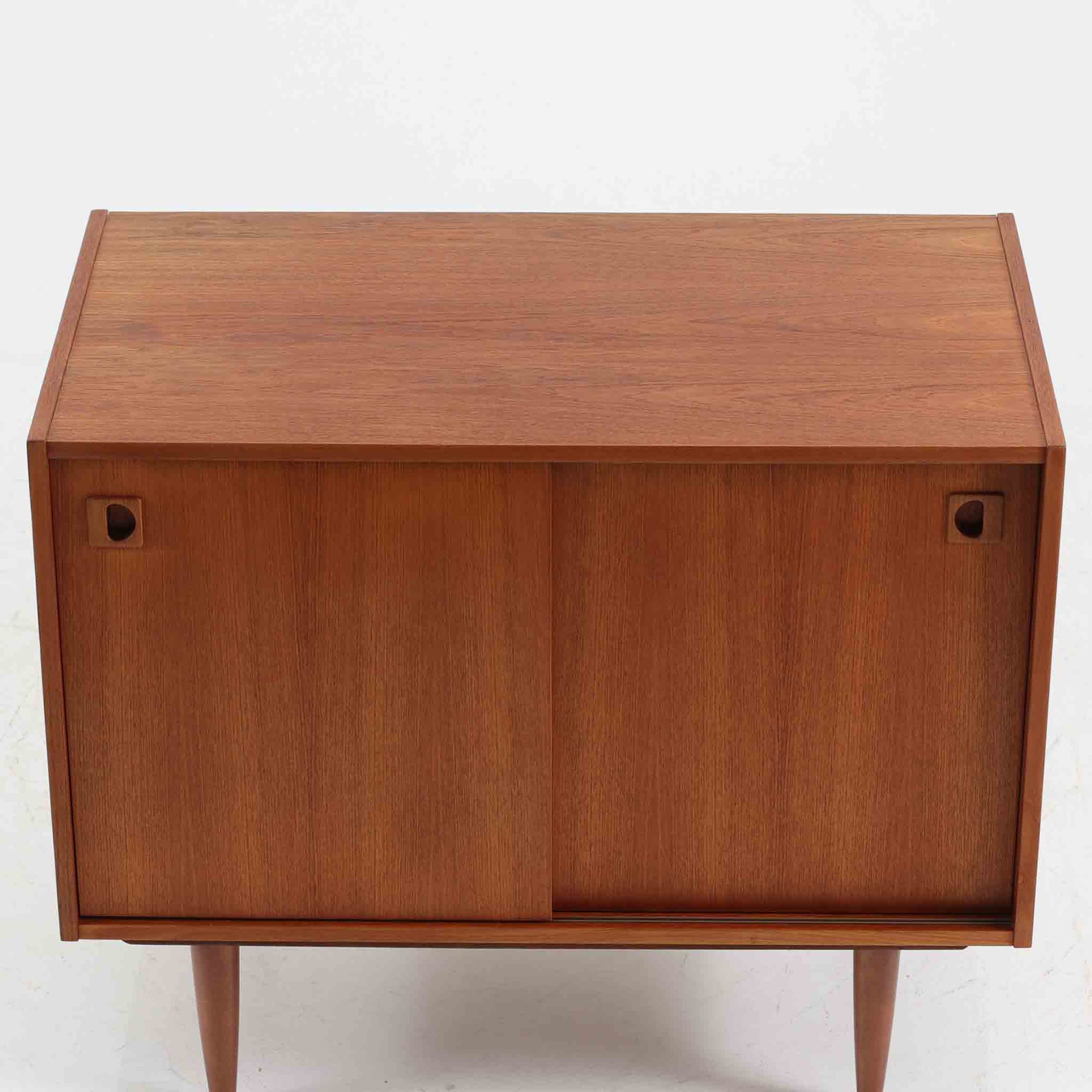 こちらの小ぶりなボードは、温かみのあるチーク材を用いたすっきりとした佇まいが魅力の北欧ビンテージ家具で、スライド扉を開くと左右に棚板付きの収納スペースが現れ、日用品や小物を効率よく整理できます。直線的な箱型の構造に、細身でテーパーの効いた脚部が軽やかな印象を添え、コンパクトながら存在感のある設計が空間に落ち着いたアクセントをもたらします。経年で深まった木肌の質感も味わい深く、リビングや玄関脇のサイド収納としても取り入れやすい一台です。