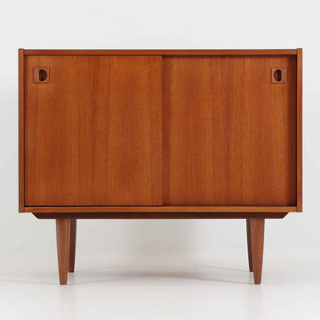 こちらの小ぶりなボードは、温かみのあるチーク材を用いたすっきりとした佇まいが魅力の北欧ビンテージ家具で、スライド扉を開くと左右に棚板付きの収納スペースが現れ、日用品や小物を効率よく整理できます。直線的な箱型の構造に、細身でテーパーの効いた脚部が軽やかな印象を添え、コンパクトながら存在感のある設計が空間に落ち着いたアクセントをもたらします。経年で深まった木肌の質感も味わい深く、リビングや玄関脇のサイド収納としても取り入れやすい一台です。