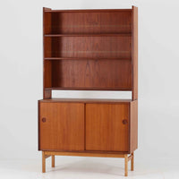 スウェーデン製のチーク材とオーク材を組み合わせたディスプレイシェルフです。幅90cmと程よいサイズ感で、上部の棚は書籍やオブジェの展示に適し、下部の引き戸収納は日用品や小物をすっきりと収めることができます。丸みを帯びた取っ手が温かみのあるアクセントとなり、北欧家具らしいシンプルで実用的なデザインを際立たせています。経年によって深みを増したチークの木目とオークのコントラストが上品な存在感を放ち、リビングや書斎の空間に落ち着いた雰囲気を添えるビンテージ家具です。