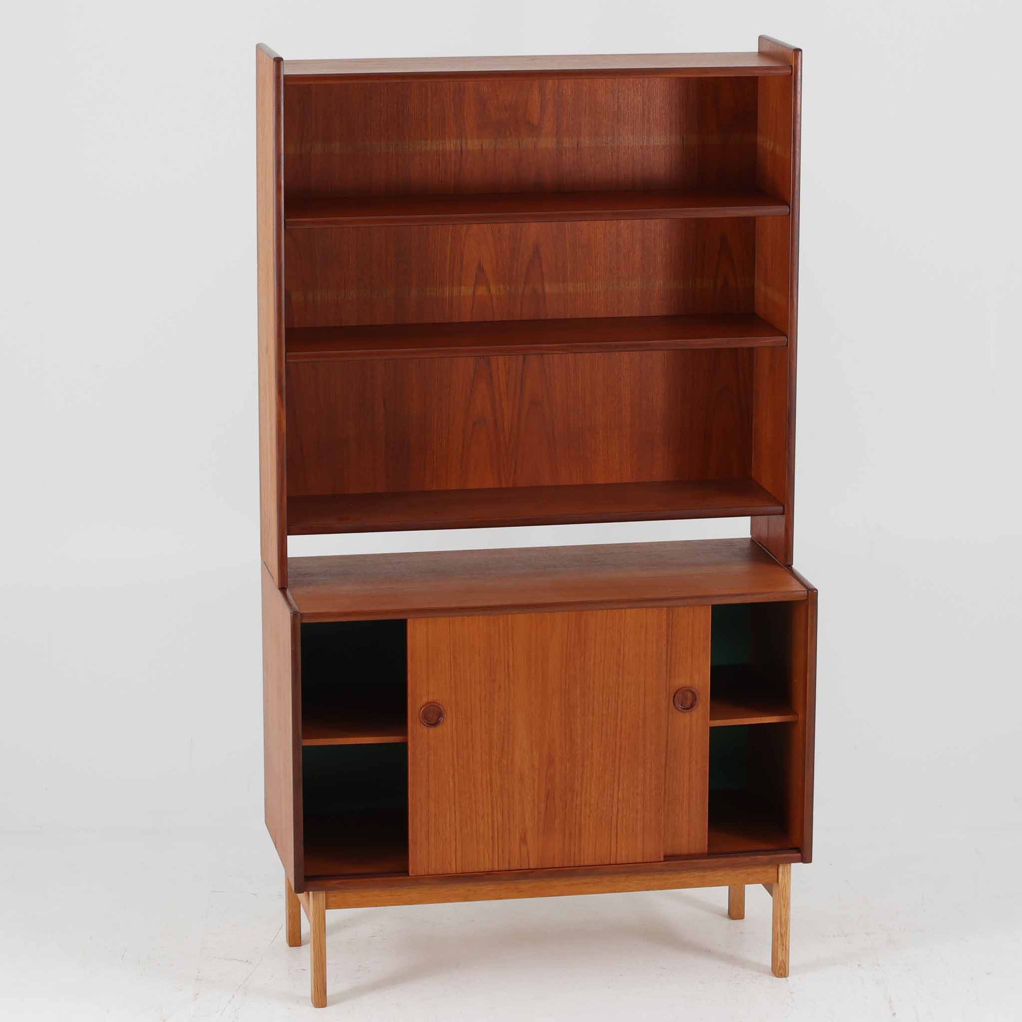 スウェーデン製のチーク材とオーク材を組み合わせたディスプレイシェルフです。幅90cmと程よいサイズ感で、上部の棚は書籍やオブジェの展示に適し、下部の引き戸収納は日用品や小物をすっきりと収めることができます。丸みを帯びた取っ手が温かみのあるアクセントとなり、北欧家具らしいシンプルで実用的なデザインを際立たせています。経年によって深みを増したチークの木目とオークのコントラストが上品な存在感を放ち、リビングや書斎の空間に落ち着いた雰囲気を添えるビンテージ家具です。