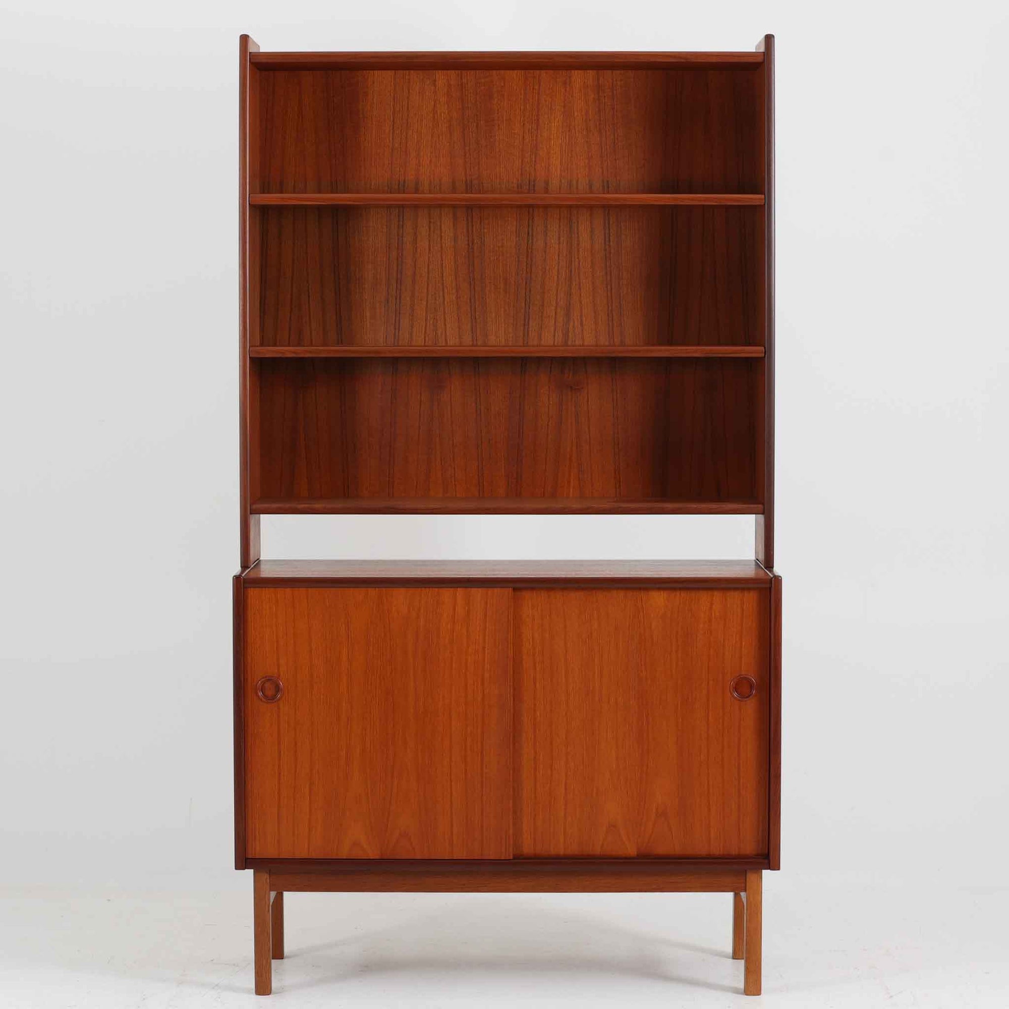 スウェーデン製のディスプレイシェルフです。上段には3段のオープンシェルフが設けられ、本や雑貨、オブジェなどを美しくディスプレイできます。下段にはスライド扉収納が備わり、内部の棚板によって整理しやすく、見せたくない収納にも便利です。チーク材の温もりある木目と、丸い掘り込み取っ手の柔らかなデザインが印象的で、北欧家具らしい穏やかな雰囲気を演出します。背面のグリーンの化粧板が空間に個性を添え、飾るアイテムをより際立たせます。本体にはチーク材、脚部にはオーク材が使用されており、素材のコントラストも魅力のひとつです。下段収納部の棚板にわずかな反りがありますが、裏返して使用することで目立たなくなります。軽やかな佇まいと実用性を兼ね備えた、北欧ビンテージらしい一台です。