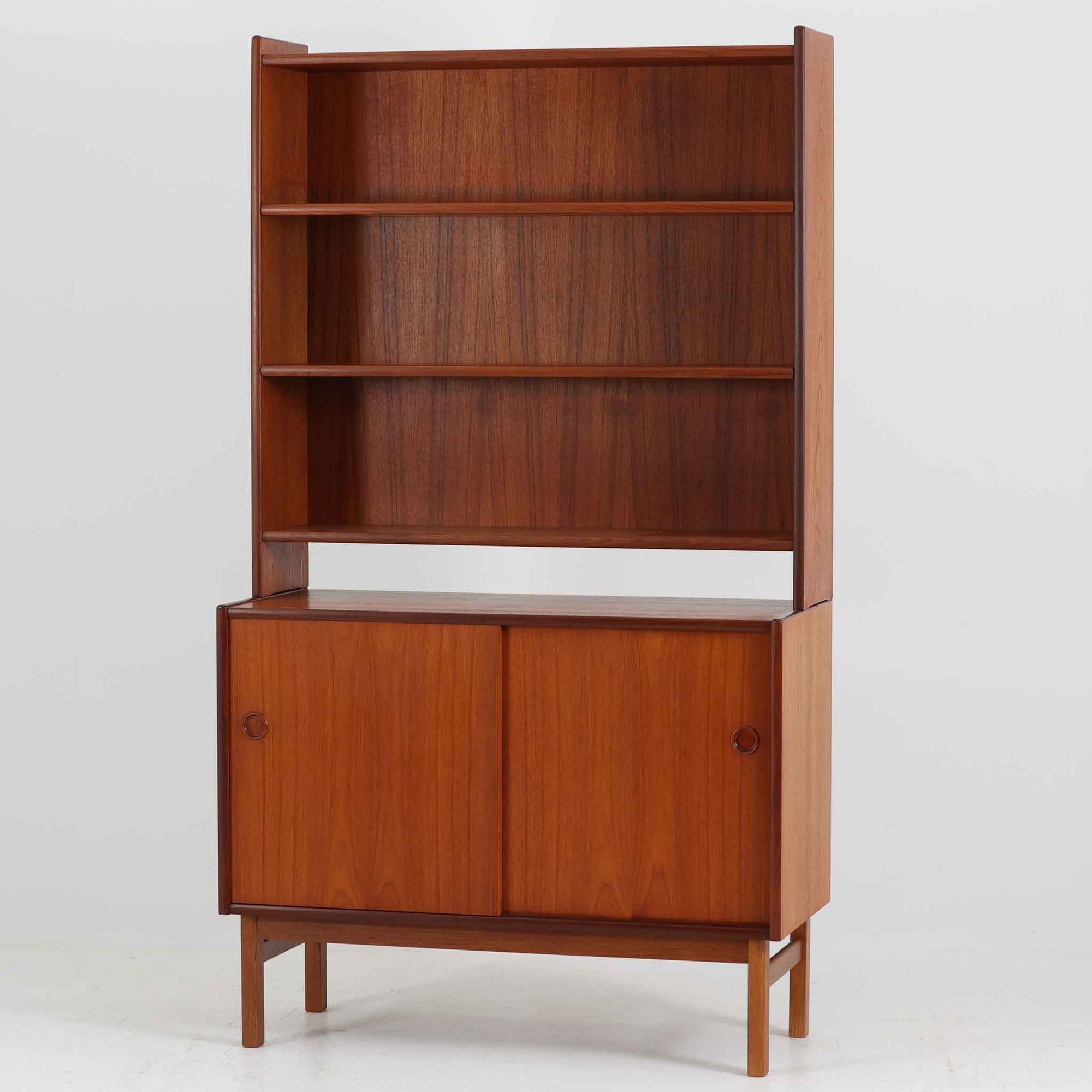 スウェーデン製のディスプレイシェルフです。上段には3段のオープンシェルフが設けられ、本や雑貨、オブジェなどを美しくディスプレイできます。下段にはスライド扉収納が備わり、内部の棚板によって整理しやすく、見せたくない収納にも便利です。チーク材の温もりある木目と、丸い掘り込み取っ手の柔らかなデザインが印象的で、北欧家具らしい穏やかな雰囲気を演出します。背面のグリーンの化粧板が空間に個性を添え、飾るアイテムをより際立たせます。本体にはチーク材、脚部にはオーク材が使用されており、素材のコントラストも魅力のひとつです。下段収納部の棚板にわずかな反りがありますが、裏返して使用することで目立たなくなります。軽やかな佇まいと実用性を兼ね備えた、北欧ビンテージらしい一台です。