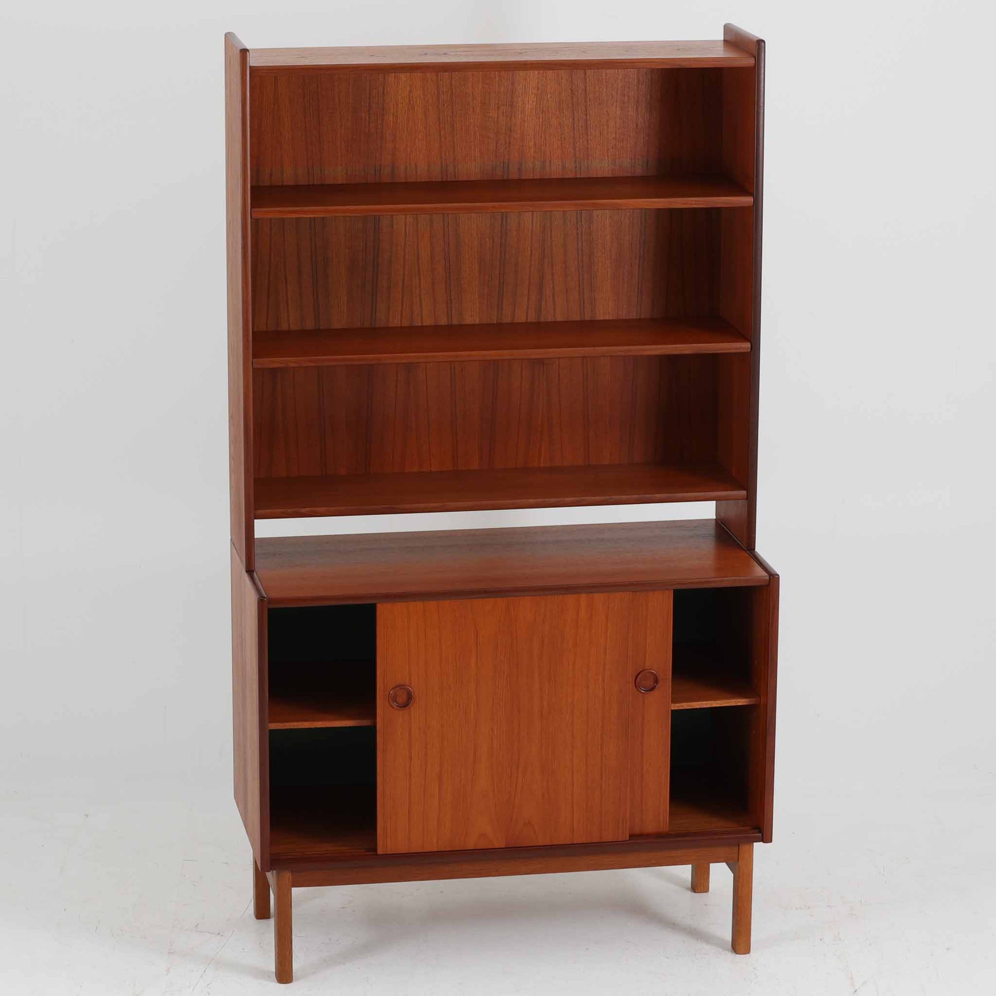 スウェーデン製のディスプレイシェルフです。上段には3段のオープンシェルフが設けられ、本や雑貨、オブジェなどを美しくディスプレイできます。下段にはスライド扉収納が備わり、内部の棚板によって整理しやすく、見せたくない収納にも便利です。チーク材の温もりある木目と、丸い掘り込み取っ手の柔らかなデザインが印象的で、北欧家具らしい穏やかな雰囲気を演出します。背面のグリーンの化粧板が空間に個性を添え、飾るアイテムをより際立たせます。本体にはチーク材、脚部にはオーク材が使用されており、素材のコントラストも魅力のひとつです。下段収納部の棚板にわずかな反りがありますが、裏返して使用することで目立たなくなります。軽やかな佇まいと実用性を兼ね備えた、北欧ビンテージらしい一台です。