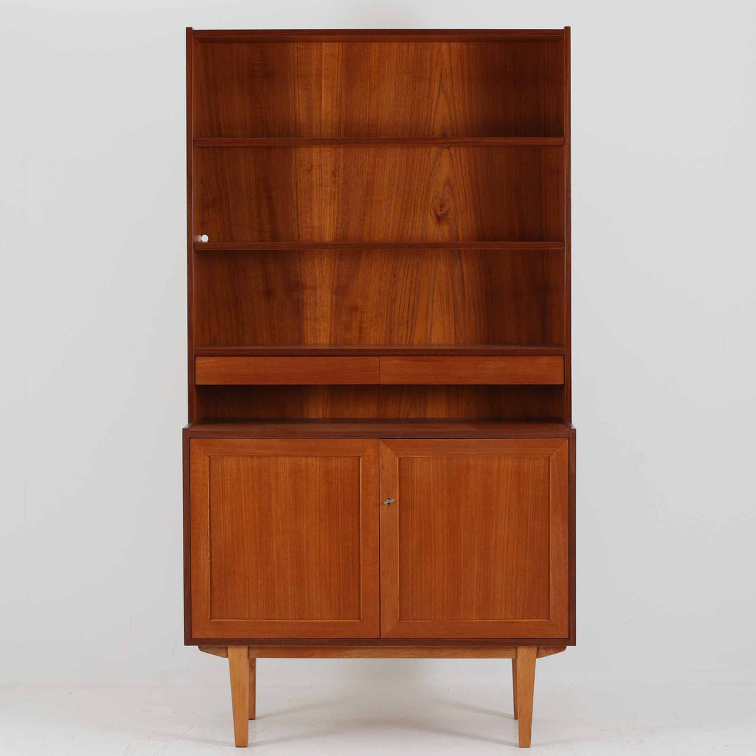 スウェーデン製のディスプレイシェルフです。上部は高さ調整可能な棚板が設けられており、書籍やオブジェの収納・展示に最適です。中央には小引き出しが2杯備わり、カトラリーや小物の整理にも便利。下部には扉付きの収納スペースがあり、日用品をすっきりと収納できます。素材にはチーク（本体）・オーク（脚の幕板）・ビーチ（脚）の3種の木材が用いられ、異なる木目のコントラストが美しい仕上がりです。北欧家具らしい温もりと実用性を兼ね備えた一台で、リビングやダイニング、書斎など幅広いシーンに調和します。