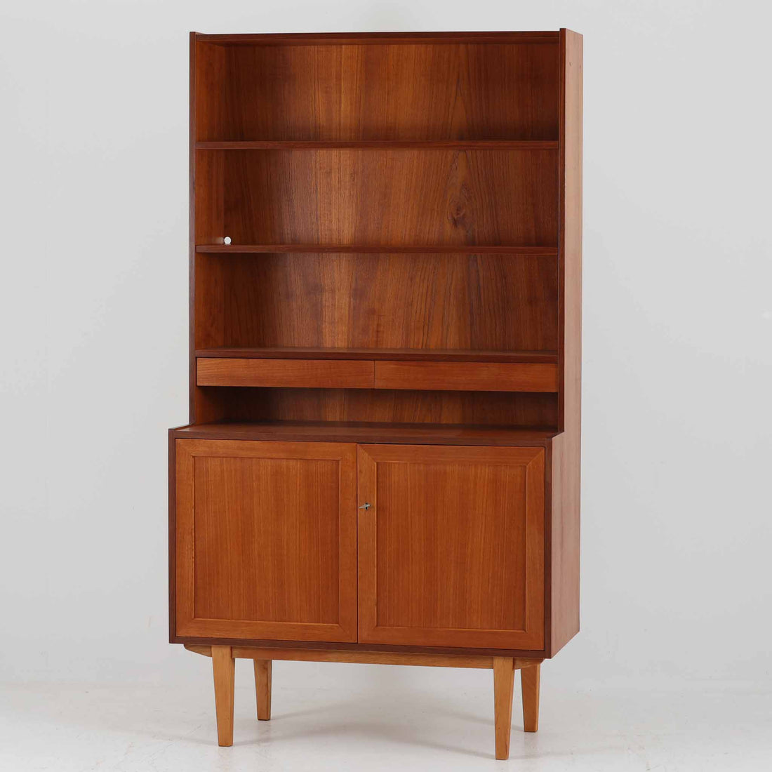 スウェーデン製のディスプレイシェルフです。上部は高さ調整可能な棚板が設けられており、書籍やオブジェの収納・展示に最適です。中央には小引き出しが2杯備わり、カトラリーや小物の整理にも便利。下部には扉付きの収納スペースがあり、日用品をすっきりと収納できます。素材にはチーク（本体）・オーク（脚の幕板）・ビーチ（脚）の3種の木材が用いられ、異なる木目のコントラストが美しい仕上がりです。北欧家具らしい温もりと実用性を兼ね備えた一台で、リビングやダイニング、書斎など幅広いシーンに調和します。