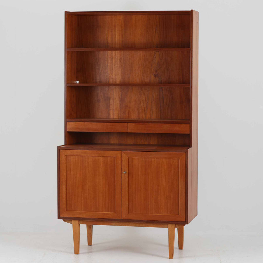 スウェーデン製のディスプレイシェルフです。上部は高さ調整可能な棚板が設けられており、書籍やオブジェの収納・展示に最適です。中央には小引き出しが2杯備わり、カトラリーや小物の整理にも便利。下部には扉付きの収納スペースがあり、日用品をすっきりと収納できます。素材にはチーク（本体）・オーク（脚の幕板）・ビーチ（脚）の3種の木材が用いられ、異なる木目のコントラストが美しい仕上がりです。北欧家具らしい温もりと実用性を兼ね備えた一台で、リビングやダイニング、書斎など幅広いシーンに調和します。