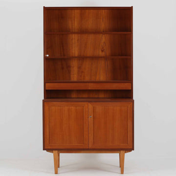 スウェーデン製のディスプレイシェルフです。上部は高さ調整可能な棚板が設けられており、書籍やオブジェの収納・展示に最適です。中央には小引き出しが2杯備わり、カトラリーや小物の整理にも便利。下部には扉付きの収納スペースがあり、日用品をすっきりと収納できます。素材にはチーク（本体）・オーク（脚の幕板）・ビーチ（脚）の3種の木材が用いられ、異なる木目のコントラストが美しい仕上がりです。北欧家具らしい温もりと実用性を兼ね備えた一台で、リビングやダイニング、書斎など幅広いシーンに調和します。