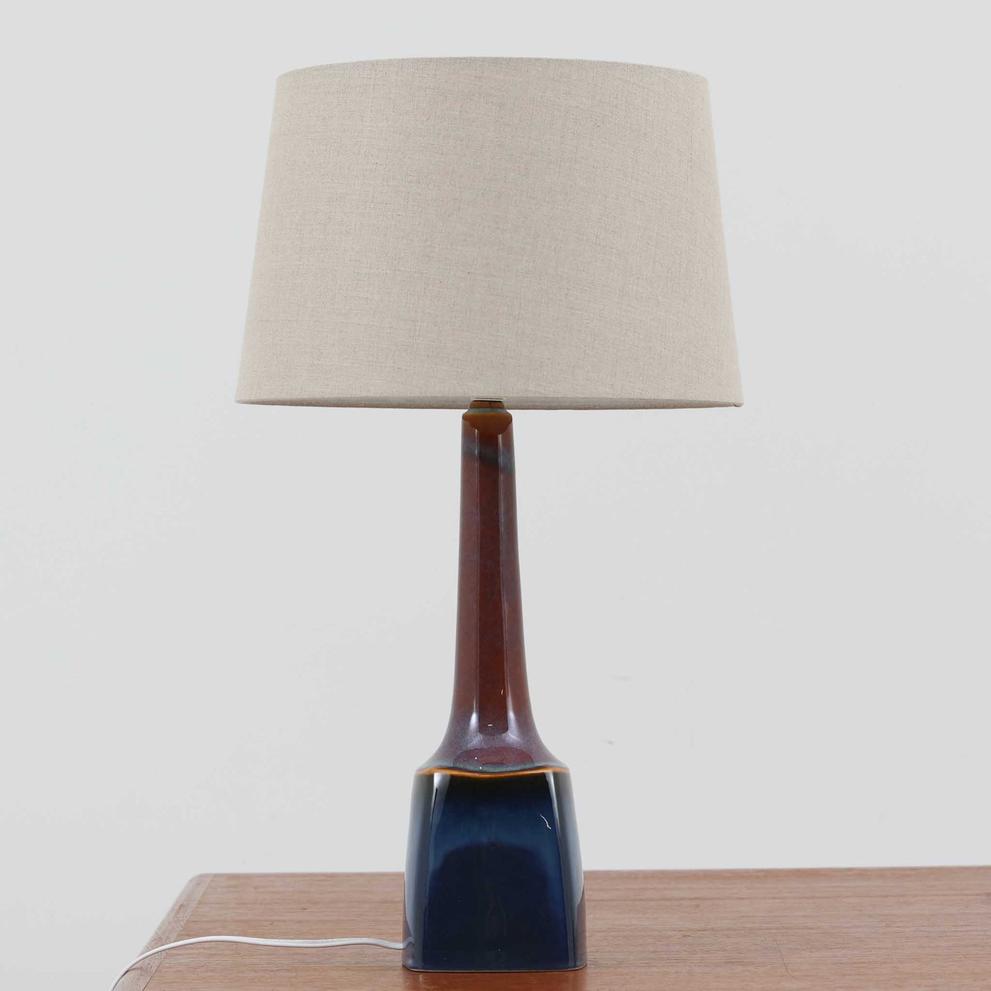 スーホルム・セラミック社製のこちらのテーブルランプは、高さ57cmと存在感のあるサイズ感が魅力の北欧ビンテージ品です。陶磁器のベースは、深みのあるブルーからブラウンへの美しい釉薬のグラデーションが特徴で、すっきりと伸びるシルエットが空間にモダンな印象を与えます。リネンシェードを透した柔らかな光はリビングや寝室を心地よく照らし、年月を経て味わいを増した質感が一点物の魅力を際立たせます。北欧インテリアのアクセントとしておすすめの逸品です。