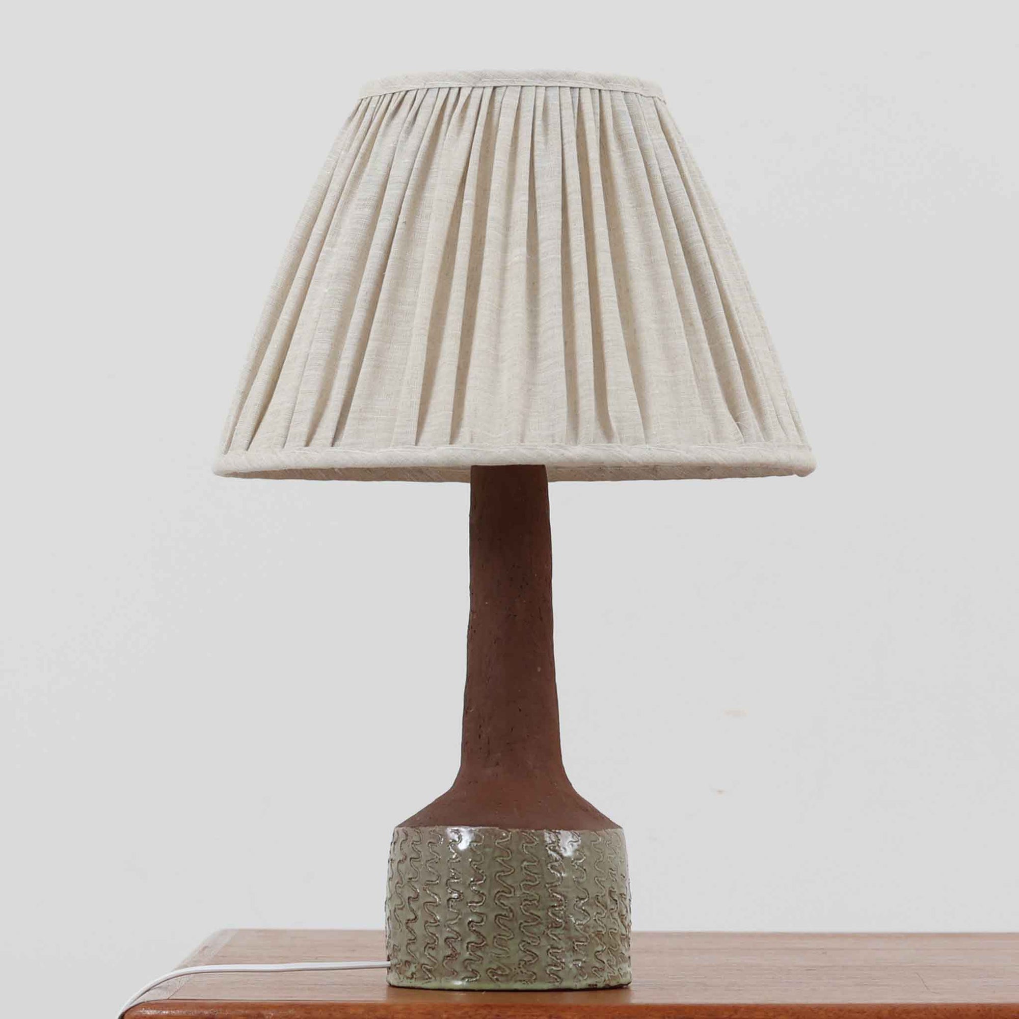 デンマーク製のテーブルランプは、高さ50cmと存在感のあるサイズが魅力の一点物ビンテージ品です。陶磁器ベースは下部に緑がかった釉薬を施し、上部は素地を生かしたマットな質感で仕上げられており、素材のコントラストがデザインの個性を際立たせています。プリーツ入りのリネンシェードを透す光はやわらかく広がり、リビングや寝室を温かみのある雰囲気に演出します。メーカー不詳・デザイナー不明ながら、Studio Potteryらしいクラフトマンシップを感じられる一点物の北欧ビンテージ品です。