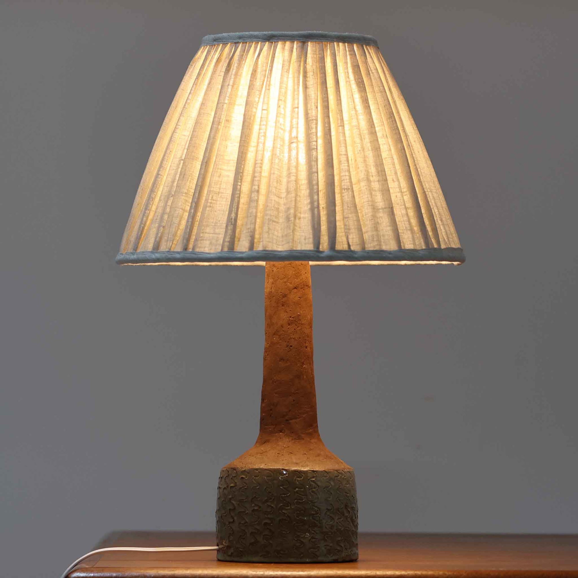 デンマーク製のテーブルランプは、高さ50cmと存在感のあるサイズが魅力の一点物ビンテージ品です。陶磁器ベースは下部に緑がかった釉薬を施し、上部は素地を生かしたマットな質感で仕上げられており、素材のコントラストがデザインの個性を際立たせています。プリーツ入りのリネンシェードを透す光はやわらかく広がり、リビングや寝室を温かみのある雰囲気に演出します。メーカー不詳・デザイナー不明ながら、Studio Potteryらしいクラフトマンシップを感じられる一点物の北欧ビンテージ品です。