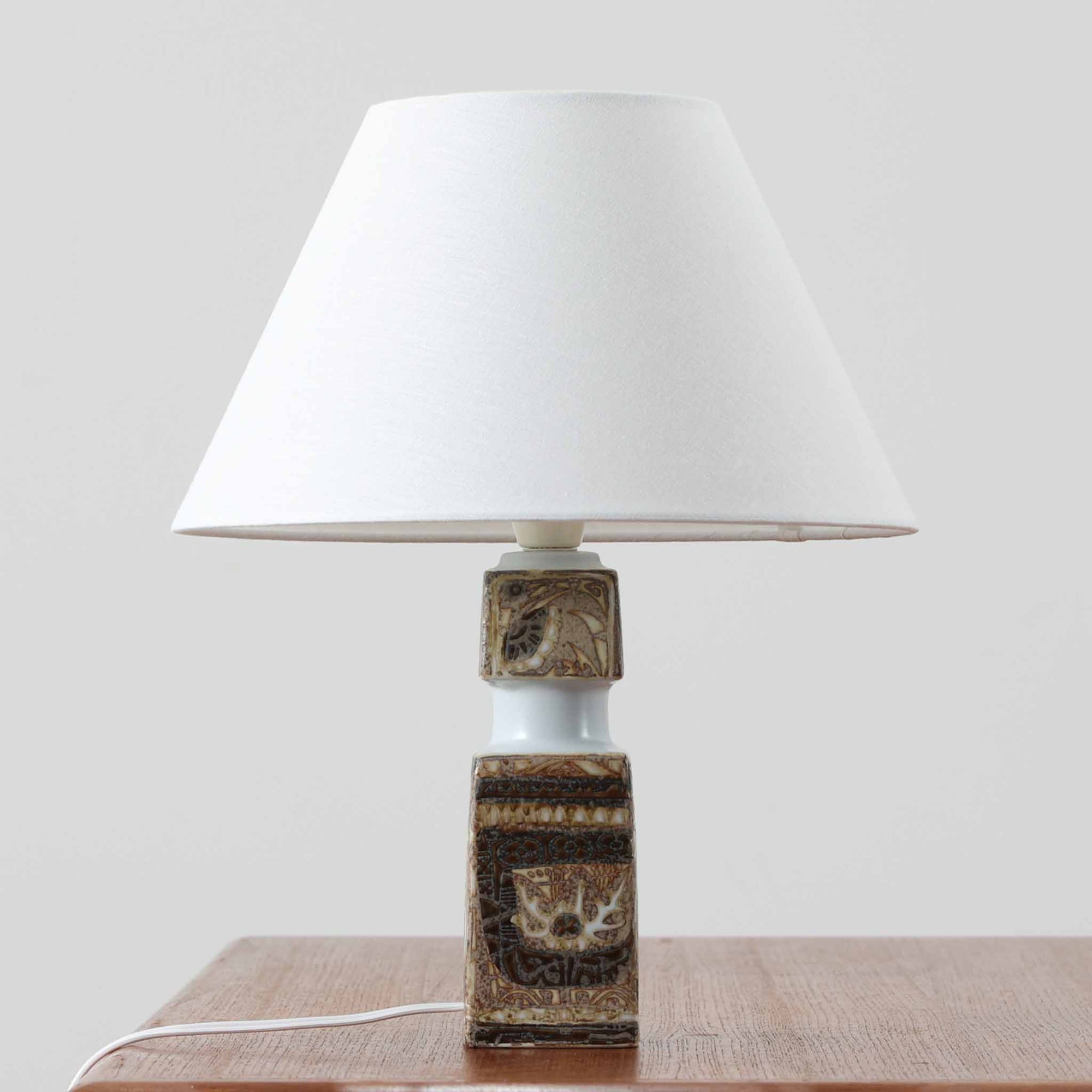 Nils Thorssonがデザインし、Royal Copenhagen社から発表された「BACA」シリーズのテーブルランプは、高さ38cmのバランスの取れたサイズ感で、北欧ビンテージらしい存在感を放ちます。陶磁器製のベースには細やかな装飾模様が施され、グラフィカルでありながら落ち着いた色調が空間に洗練された雰囲気を添えます。リネンシェードを透過する柔らかな光が温もりを演出し、リビングやベッドサイドに最適です。年月を経て深みを増した質感と工芸的な美しさを楽しめる一点物です。