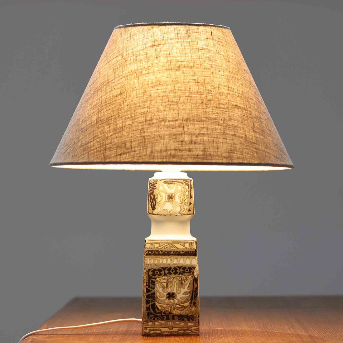 Nils Thorssonがデザインし、Royal Copenhagen社から発表された「BACA」シリーズのテーブルランプは、高さ38cmのバランスの取れたサイズ感で、北欧ビンテージらしい存在感を放ちます。陶磁器製のベースには細やかな装飾模様が施され、グラフィカルでありながら落ち着いた色調が空間に洗練された雰囲気を添えます。リネンシェードを透過する柔らかな光が温もりを演出し、リビングやベッドサイドに最適です。年月を経て深みを増した質感と工芸的な美しさを楽しめる一点物です。
