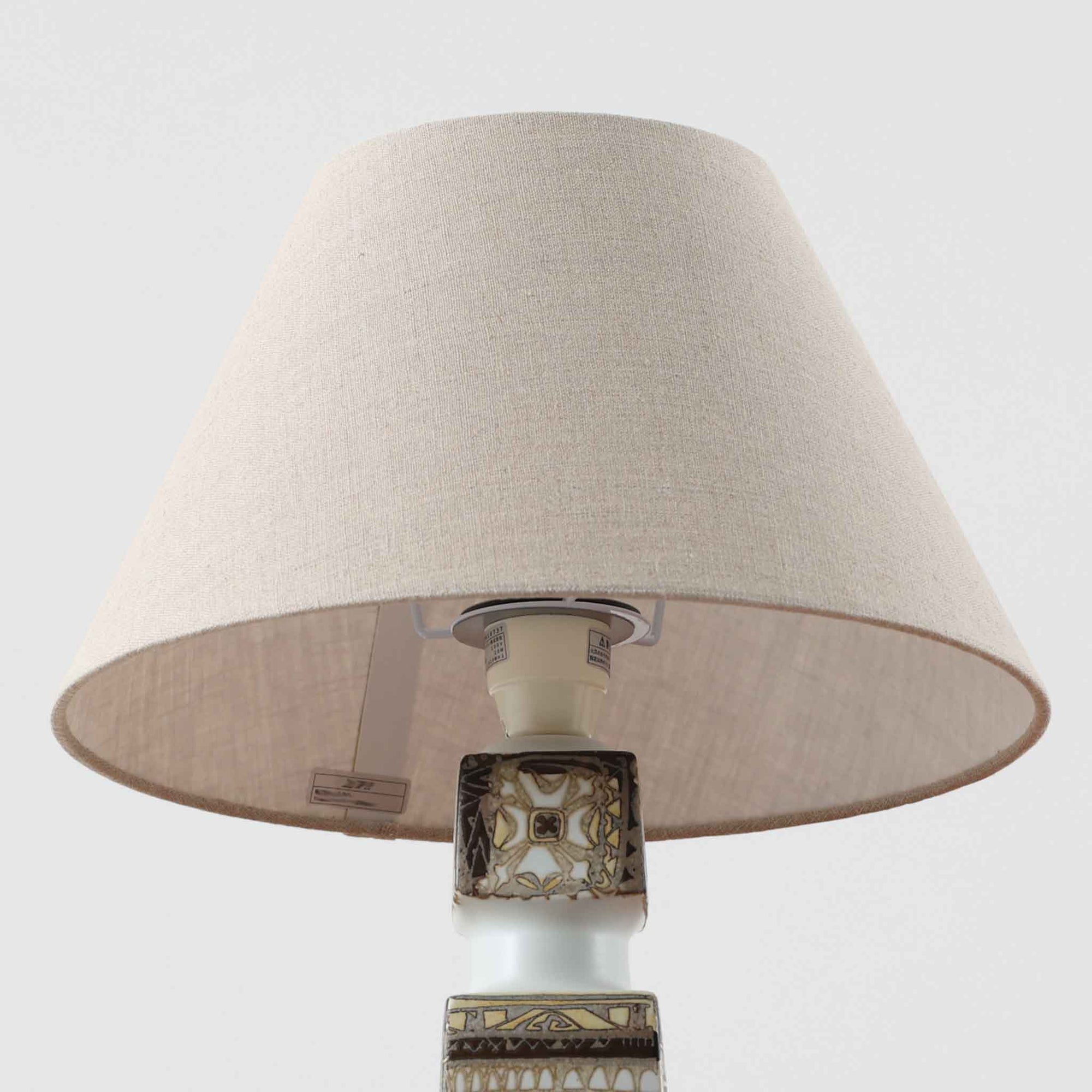 Nils Thorssonがデザインし、Royal Copenhagen社から発表された「BACA」シリーズのテーブルランプは、高さ38cmのバランスの取れたサイズ感で、北欧ビンテージらしい存在感を放ちます。陶磁器製のベースには細やかな装飾模様が施され、グラフィカルでありながら落ち着いた色調が空間に洗練された雰囲気を添えます。リネンシェードを透過する柔らかな光が温もりを演出し、リビングやベッドサイドに最適です。年月を経て深みを増した質感と工芸的な美しさを楽しめる一点物です。