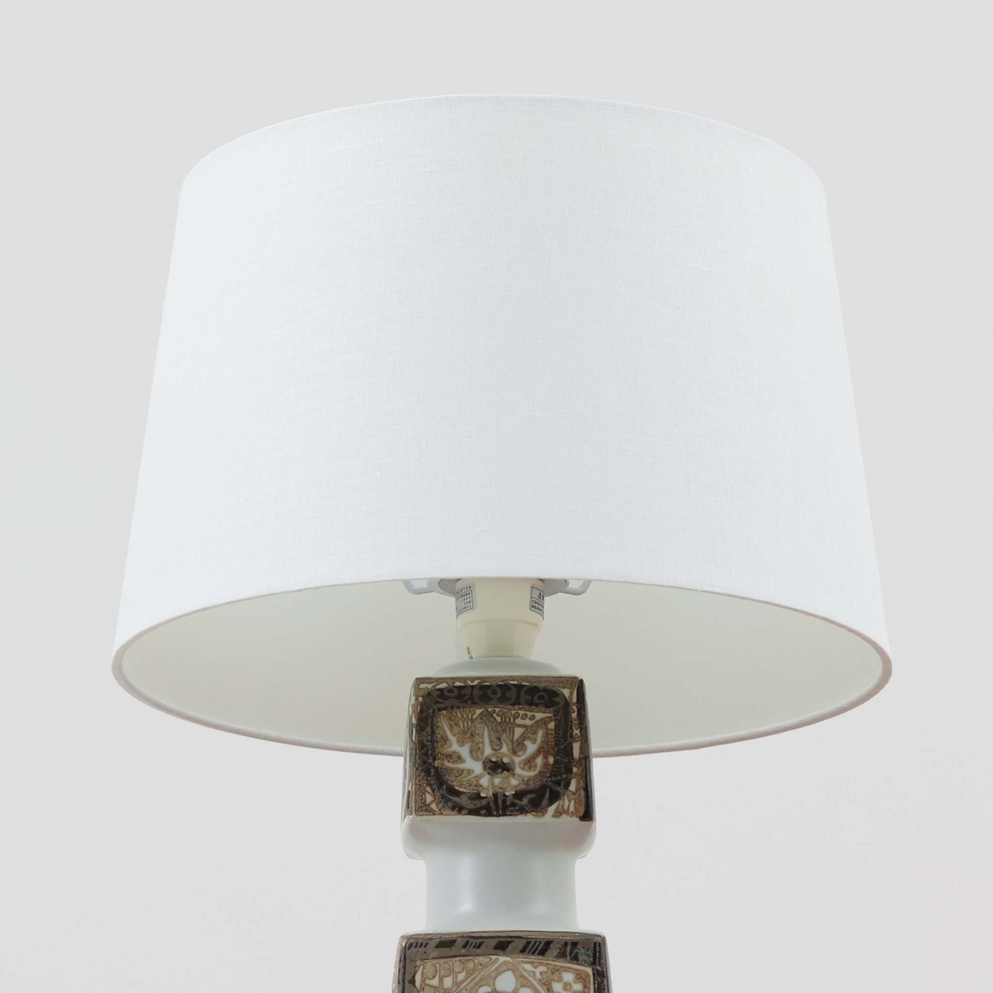 Nils Thorssonがデザインし、Royal Copenhagen社から発表されたBACAシリーズのこちらのテーブルランプは、高さ50cmと大きめのサイズで空間に存在感を放ちます。陶磁器製のベースにはトールソンならではの幾何学的かつ装飾的なパターンが施され、芸術性とクラフト感が融合した独特の魅力を漂わせています。リネンシェードを通した柔らかな光はリビングやベッドサイドに落ち着きをもたらし、ビンテージならではの経年の深みが一点物としての価値を高めています。