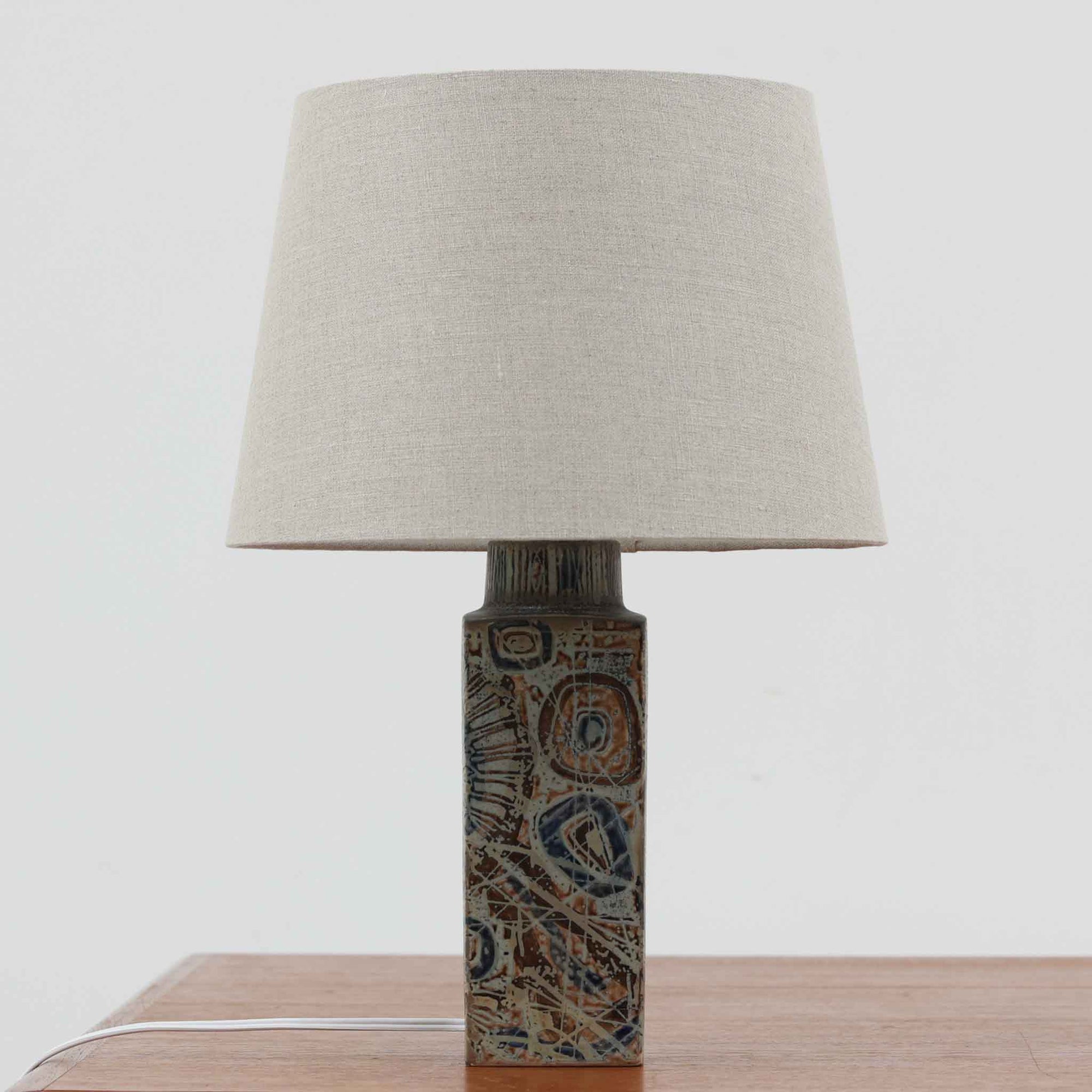 Nils Thorssonが手掛けたRoyal Copenhagen社BACAシリーズのテーブルランプは、高さ41cmの程よいサイズ感で空間に上質な存在感を与えます。陶磁器のベースには幾何学的で抽象的なモチーフが施され、複雑な釉薬の表情が芸術性を引き立てています。シンプルなリネンシェードを透した光は柔らかく広がり、リビングやベッドサイドを温かみのある雰囲気に演出。クラフトマンシップとデザイン性が融合した北欧ビンテージの魅力を存分に味わえる逸品です。