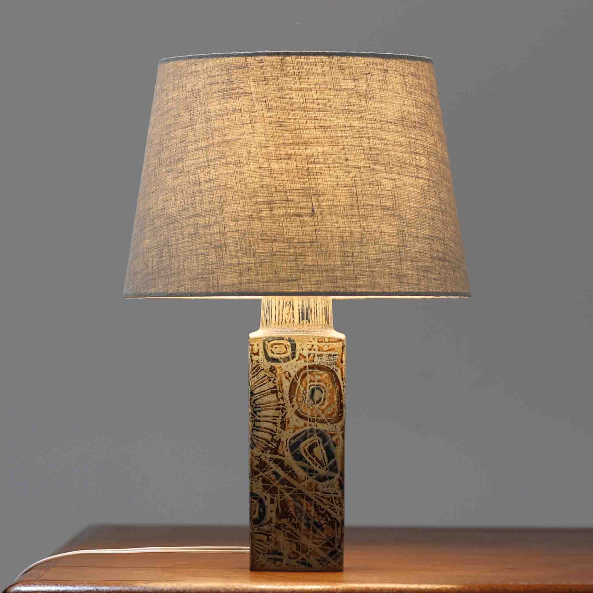 Nils Thorssonが手掛けたRoyal Copenhagen社BACAシリーズのテーブルランプは、高さ41cmの程よいサイズ感で空間に上質な存在感を与えます。陶磁器のベースには幾何学的で抽象的なモチーフが施され、複雑な釉薬の表情が芸術性を引き立てています。シンプルなリネンシェードを透した光は柔らかく広がり、リビングやベッドサイドを温かみのある雰囲気に演出。クラフトマンシップとデザイン性が融合した北欧ビンテージの魅力を存分に味わえる逸品です。