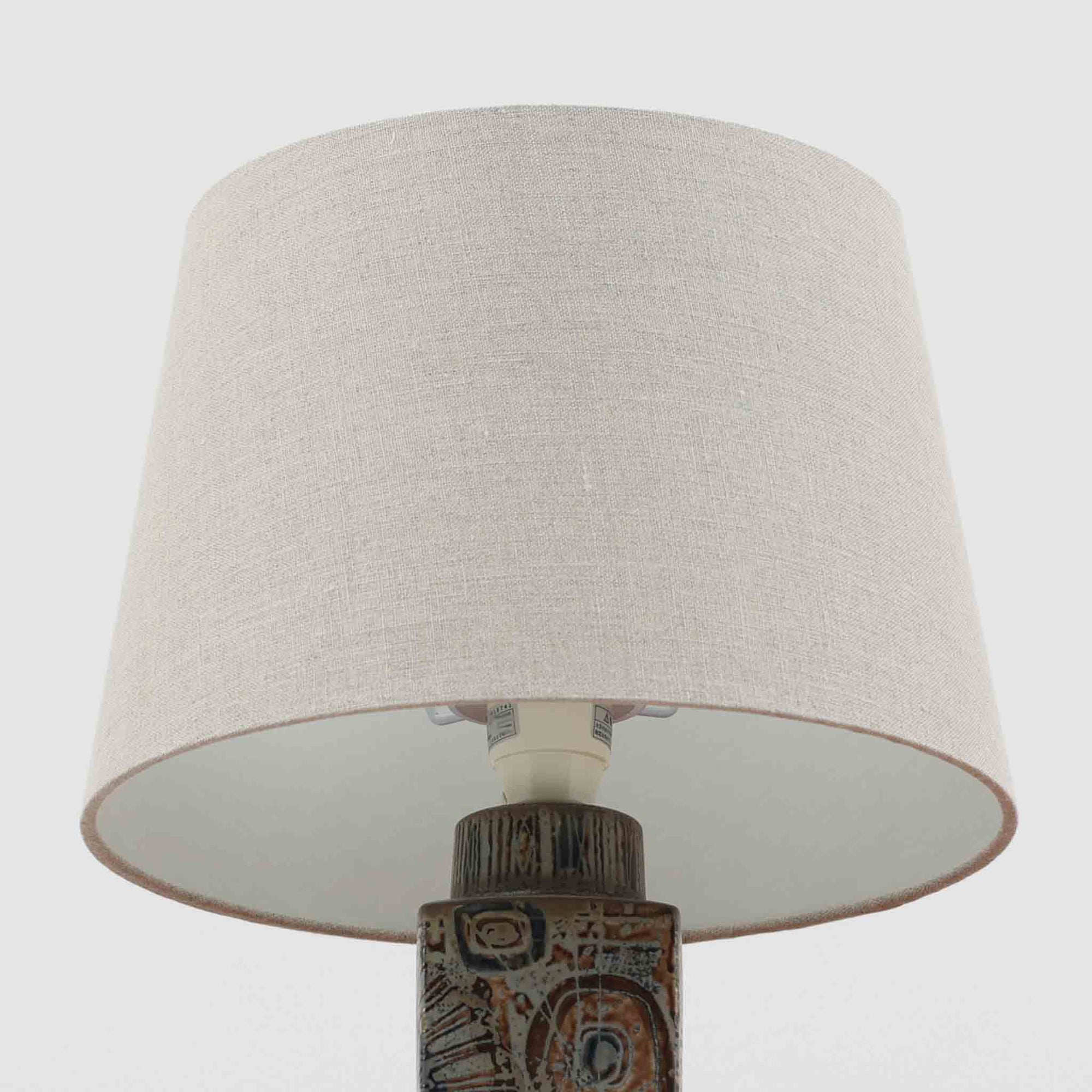 Nils Thorssonが手掛けたRoyal Copenhagen社BACAシリーズのテーブルランプは、高さ41cmの程よいサイズ感で空間に上質な存在感を与えます。陶磁器のベースには幾何学的で抽象的なモチーフが施され、複雑な釉薬の表情が芸術性を引き立てています。シンプルなリネンシェードを透した光は柔らかく広がり、リビングやベッドサイドを温かみのある雰囲気に演出。クラフトマンシップとデザイン性が融合した北欧ビンテージの魅力を存分に味わえる逸品です。