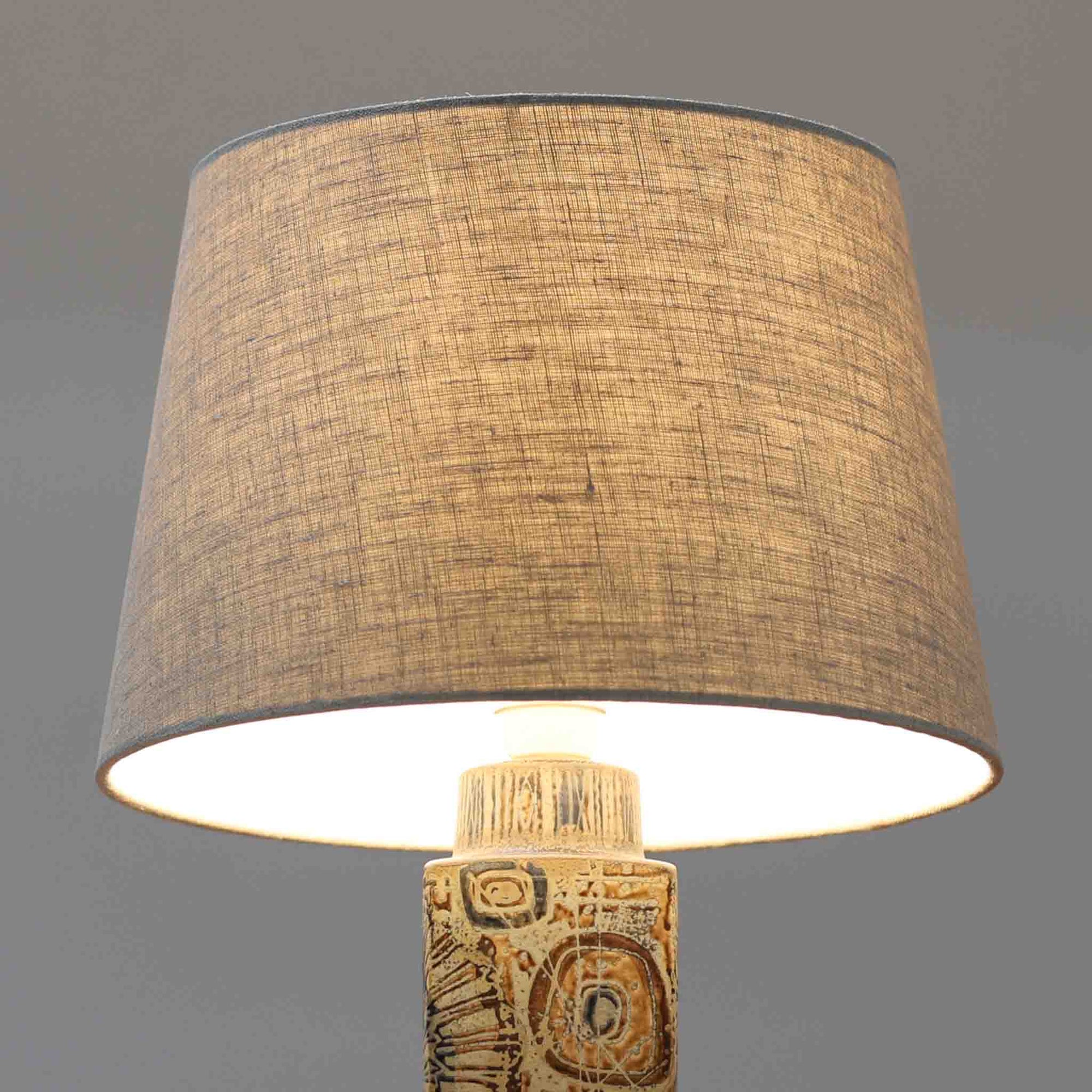 Nils Thorssonが手掛けたRoyal Copenhagen社BACAシリーズのテーブルランプは、高さ41cmの程よいサイズ感で空間に上質な存在感を与えます。陶磁器のベースには幾何学的で抽象的なモチーフが施され、複雑な釉薬の表情が芸術性を引き立てています。シンプルなリネンシェードを透した光は柔らかく広がり、リビングやベッドサイドを温かみのある雰囲気に演出。クラフトマンシップとデザイン性が融合した北欧ビンテージの魅力を存分に味わえる逸品です。