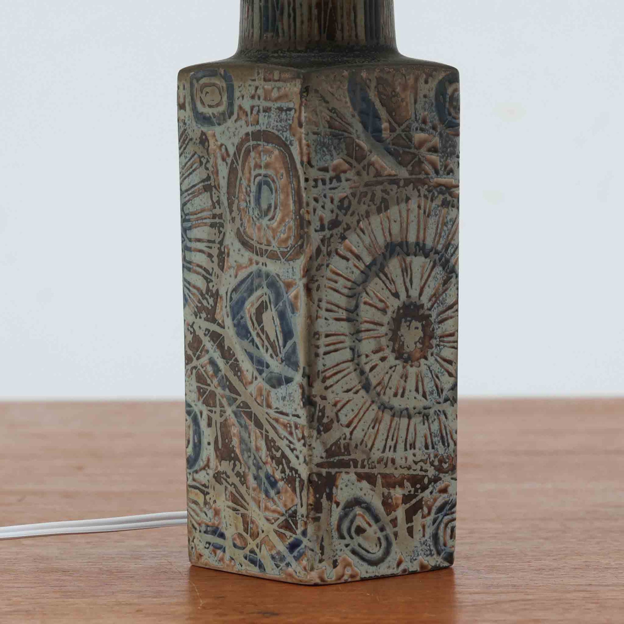 Nils Thorssonが手掛けたRoyal Copenhagen社BACAシリーズのテーブルランプは、高さ41cmの程よいサイズ感で空間に上質な存在感を与えます。陶磁器のベースには幾何学的で抽象的なモチーフが施され、複雑な釉薬の表情が芸術性を引き立てています。シンプルなリネンシェードを透した光は柔らかく広がり、リビングやベッドサイドを温かみのある雰囲気に演出。クラフトマンシップとデザイン性が融合した北欧ビンテージの魅力を存分に味わえる逸品です。