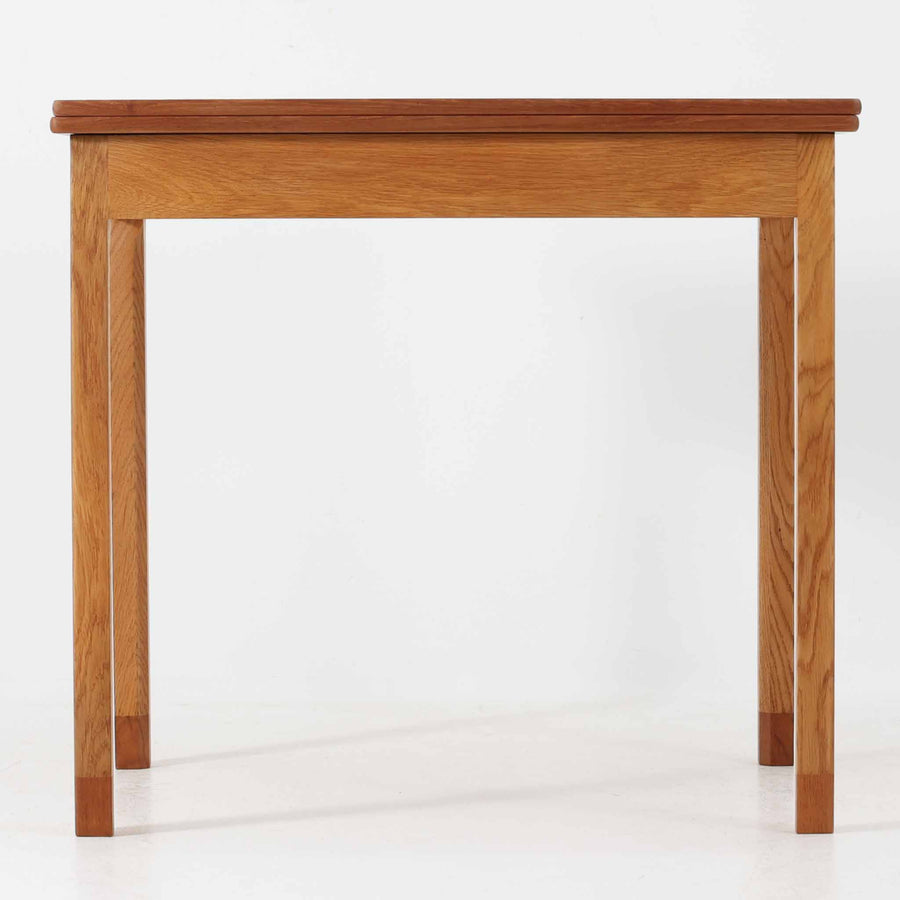 Borge Mogensen（ボーエ・モーエンセン）がSoborgのために手掛けた「Folding table」は、チーク材の天板とオーク材の脚部を組み合わせた温かみある佇まいが魅力のダイニングテーブルで、中央の蝶番を開閉して幅80～120cmへと柔軟に調整できる構造が特徴です。内部にはモーエンセン家具らしい実用性を備えた収納スペースが設けられ、ゲームテーブルとして親しまれてきた背景を感じさせます。経年により深まった木肌の風合いがビンテージならではの味わいをまとい、ダイニングやワークスペースのサブテーブルとしても多用途に活躍する一台です。また、裏面には「SKM」の刻印が確認でき、スウェーデン軍（Svenska KrigsMakten）の備品として使用されていたことを示しています。