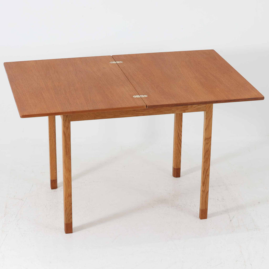 Borge Mogensen（ボーエ・モーエンセン）がSoborgのために手掛けた「Folding table」は、チーク材の天板とオーク材の脚部を組み合わせた温かみある佇まいが魅力のダイニングテーブルで、中央の蝶番を開閉して幅80～120cmへと柔軟に調整できる構造が特徴です。内部にはモーエンセン家具らしい実用性を備えた収納スペースが設けられ、ゲームテーブルとして親しまれてきた背景を感じさせます。経年により深まった木肌の風合いがビンテージならではの味わいをまとい、ダイニングやワークスペースのサブテーブルとしても多用途に活躍する一台です。また、裏面には「SKM」の刻印が確認でき、スウェーデン軍（Svenska KrigsMakten）の備品として使用されていたことを示しています。