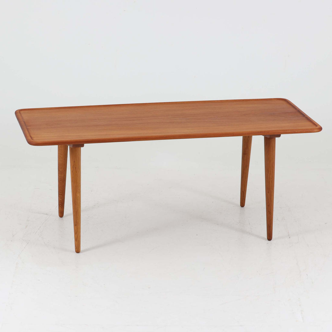 Hans J. Wegner（ハンス・J・ウェグナー）によるAndreas Tuck社製のセンターテーブル「AT11」です。幅130cmの広々とした天板はチーク無垢材を使用し、縁には繊細な面取り加工が施され、柔らかな印象を与えます。脚部には堅牢なオーク無垢材が用いられ、軽やかなテーパードデザインが北欧家具らしいバランスの良さを感じさせます。さらに脚のネジ部分まで木製で仕上げられており、細部に至るまで職人技が光る逸品です。脚は取り外し可能な構造で、輸送や設置も容易です。シンプルでありながら素材の美しさが際立ち、経年変化による深みを増した木肌がビンテージならではの存在感を放ちます。リビングの中心に置いて使うことで、空間に上質な雰囲気を添える一台です。