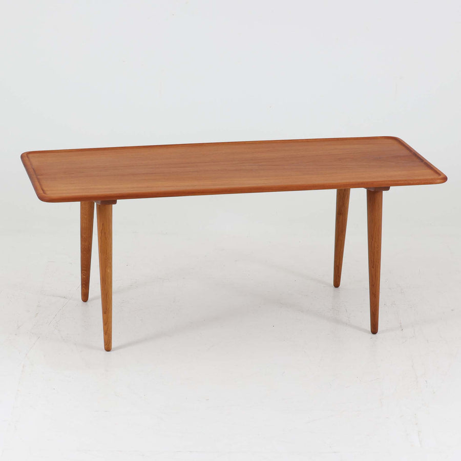 Hans J. Wegner（ハンス・J・ウェグナー）によるAndreas Tuck社製のセンターテーブル「AT11」です。幅130cmの広々とした天板はチーク無垢材を使用し、縁には繊細な面取り加工が施され、柔らかな印象を与えます。脚部には堅牢なオーク無垢材が用いられ、軽やかなテーパードデザインが北欧家具らしいバランスの良さを感じさせます。さらに脚のネジ部分まで木製で仕上げられており、細部に至るまで職人技が光る逸品です。脚は取り外し可能な構造で、輸送や設置も容易です。シンプルでありながら素材の美しさが際立ち、経年変化による深みを増した木肌がビンテージならではの存在感を放ちます。リビングの中心に置いて使うことで、空間に上質な雰囲気を添える一台です。