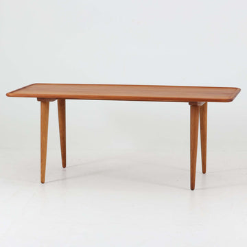 Hans J. Wegner（ハンス・J・ウェグナー）によるAndreas Tuck社製のセンターテーブル「AT11」です。幅130cmの広々とした天板はチーク無垢材を使用し、縁には繊細な面取り加工が施され、柔らかな印象を与えます。脚部には堅牢なオーク無垢材が用いられ、軽やかなテーパードデザインが北欧家具らしいバランスの良さを感じさせます。さらに脚のネジ部分まで木製で仕上げられており、細部に至るまで職人技が光る逸品です。脚は取り外し可能な構造で、輸送や設置も容易です。シンプルでありながら素材の美しさが際立ち、経年変化による深みを増した木肌がビンテージならではの存在感を放ちます。リビングの中心に置いて使うことで、空間に上質な雰囲気を添える一台です。